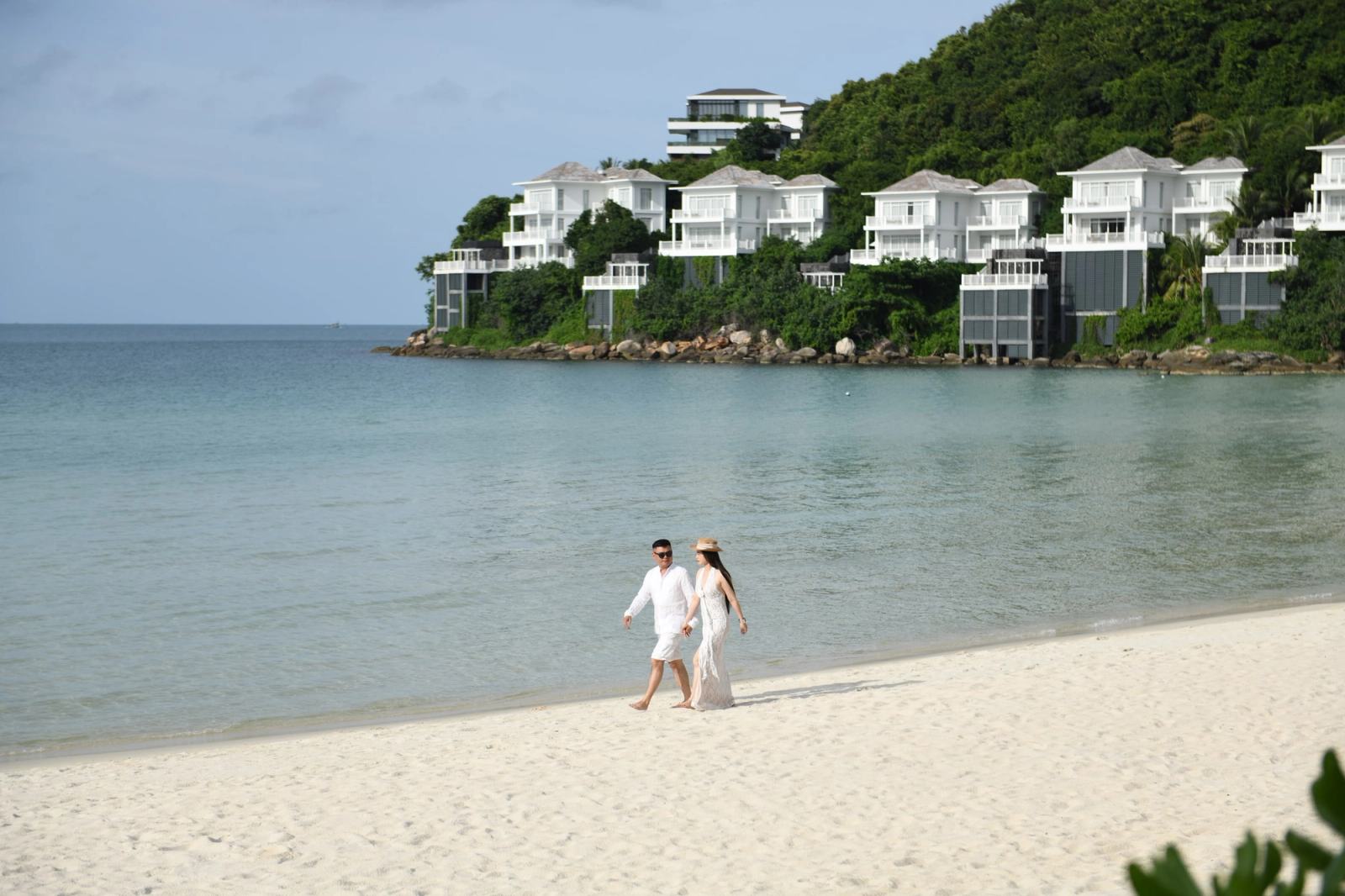 Couple walking along a sandy beach with white hillside villas across the bay.