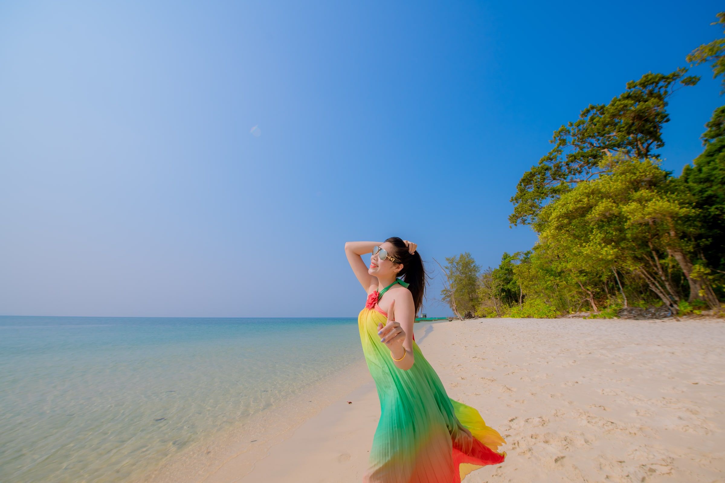 Woman in a colourful dress standing on a sandy beach beside turquoise water.
