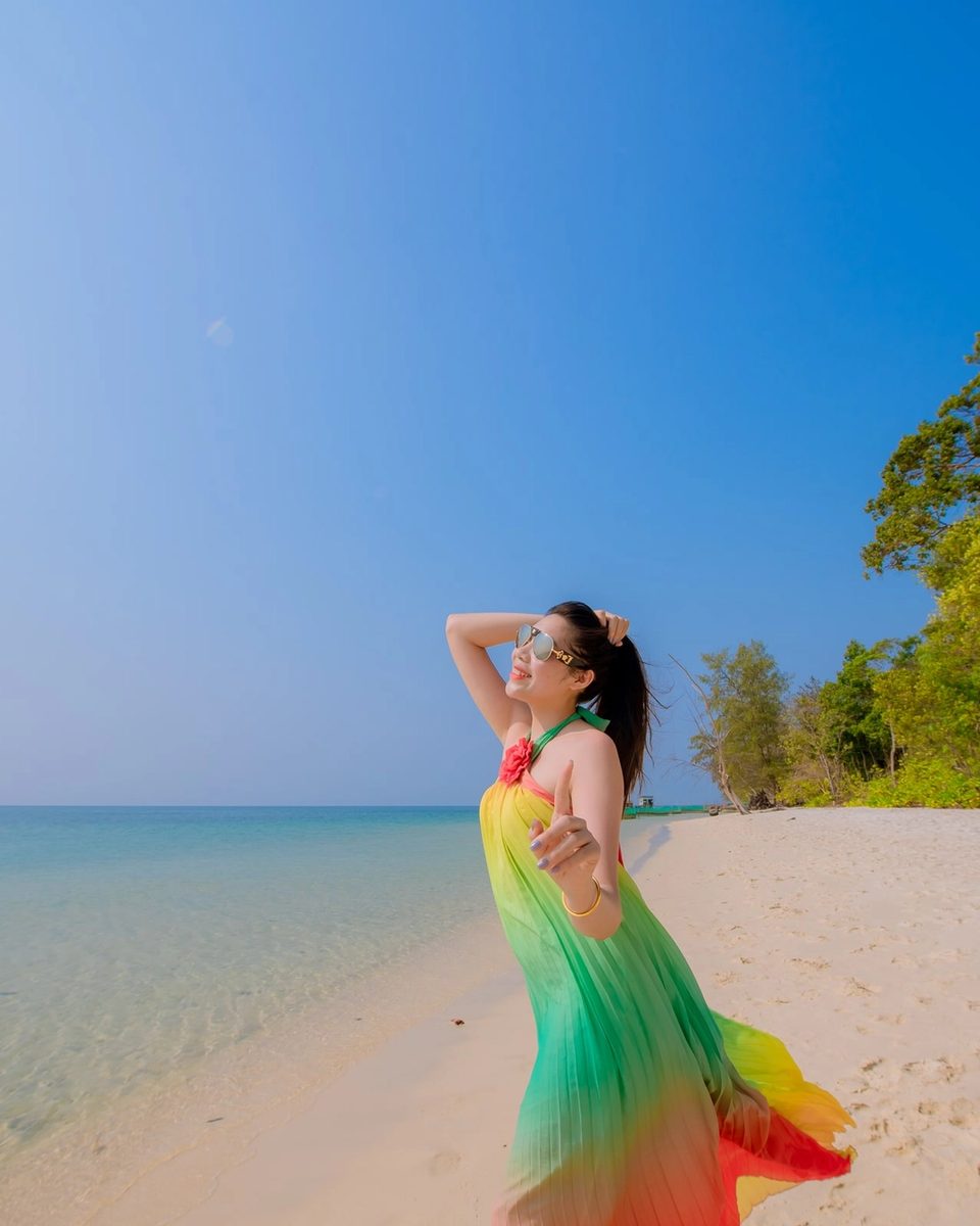 Woman in a colourful dress standing on a sandy beach beside turquoise water.