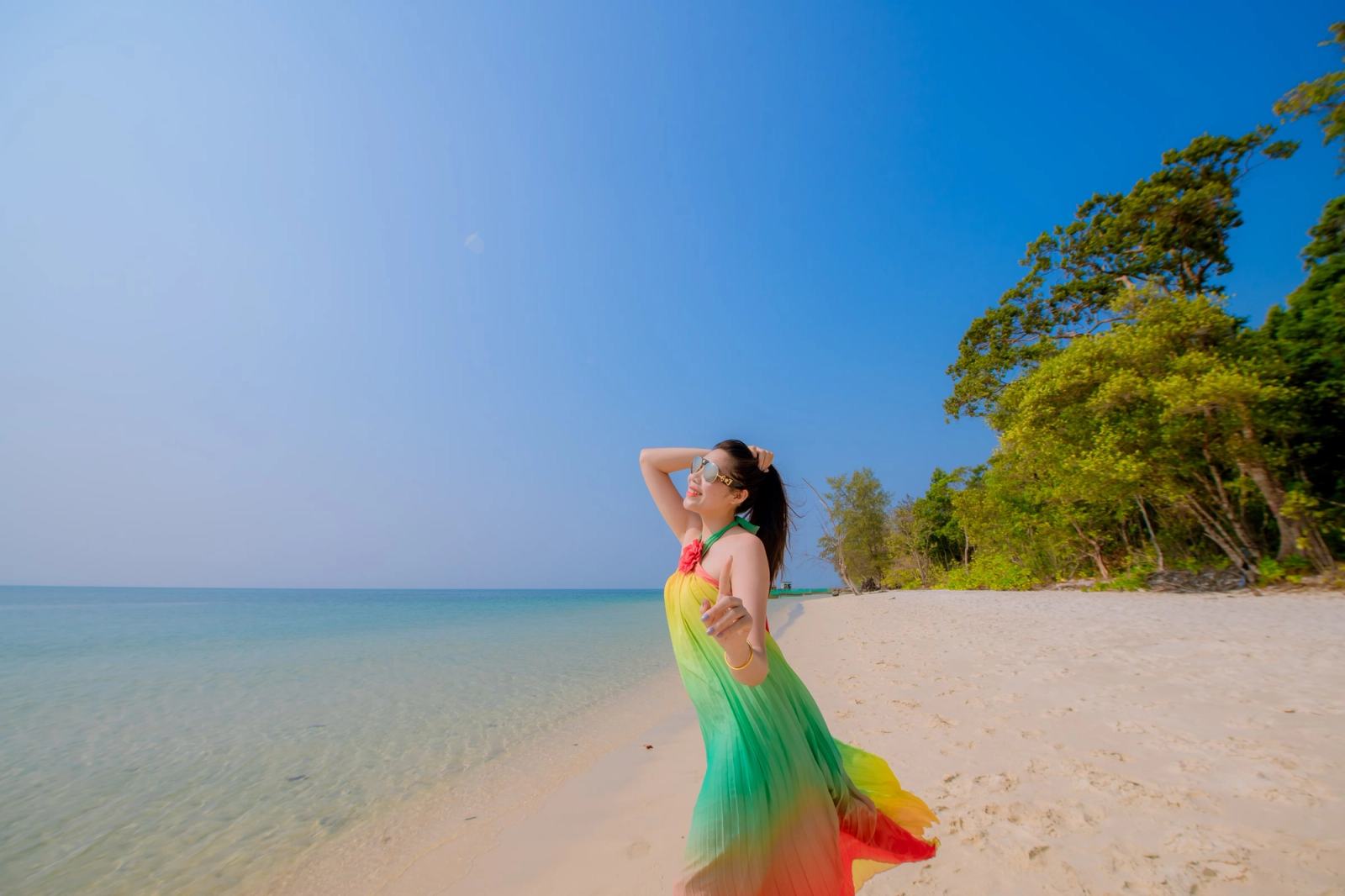 Woman in a colourful dress standing on a sandy beach beside turquoise water.