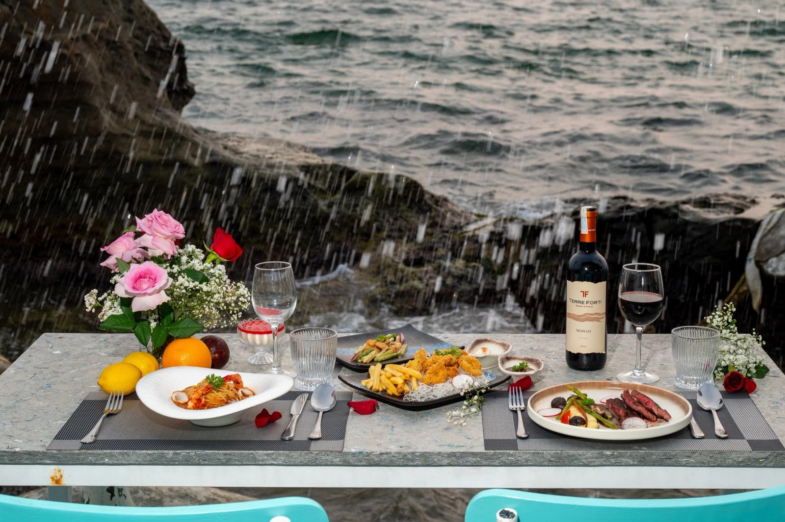 Dishes, wine and flowers on a seaside table with a wave splashing against rocks behind it.
