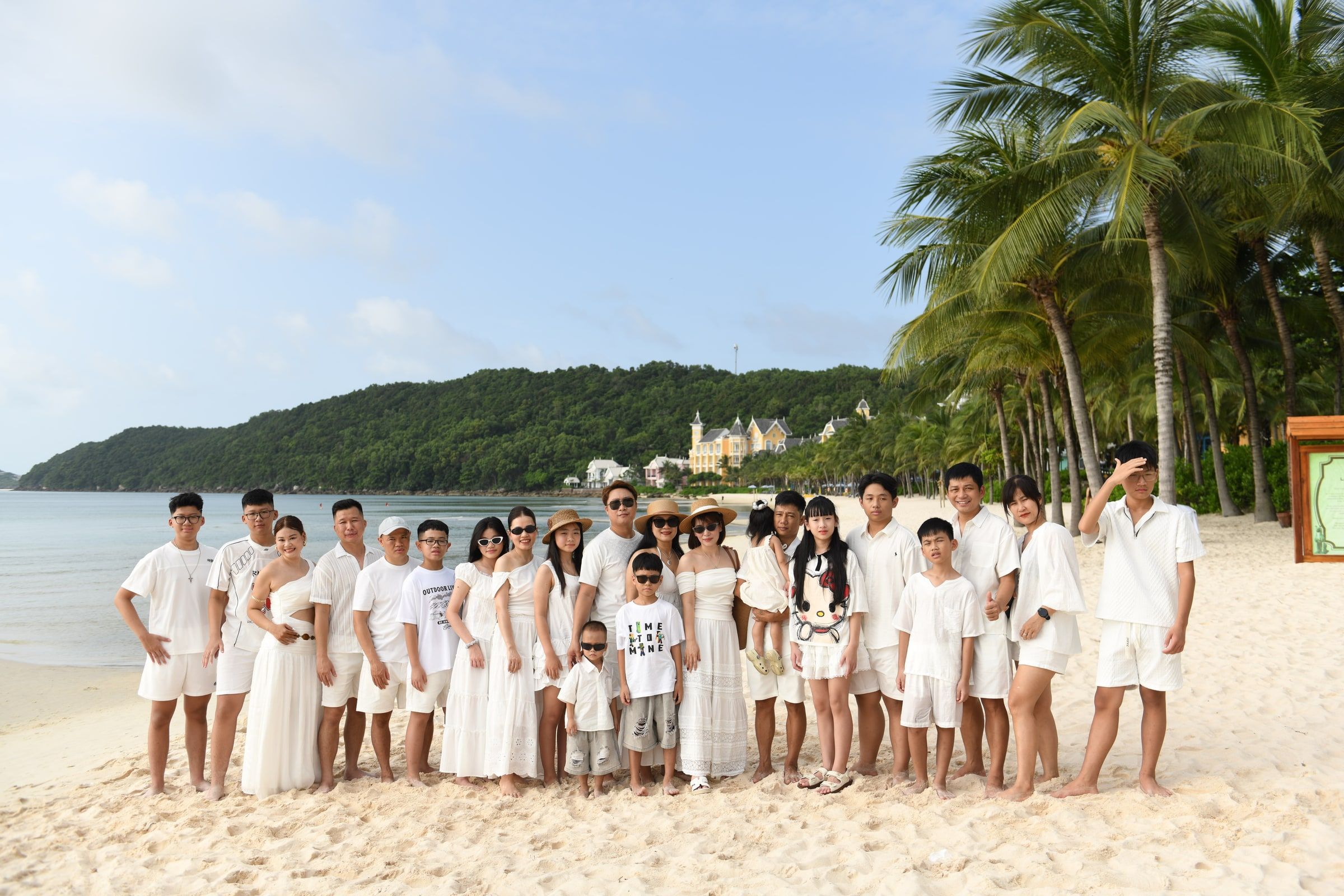 Large family group in white outfits posing on a beach beside palm trees.