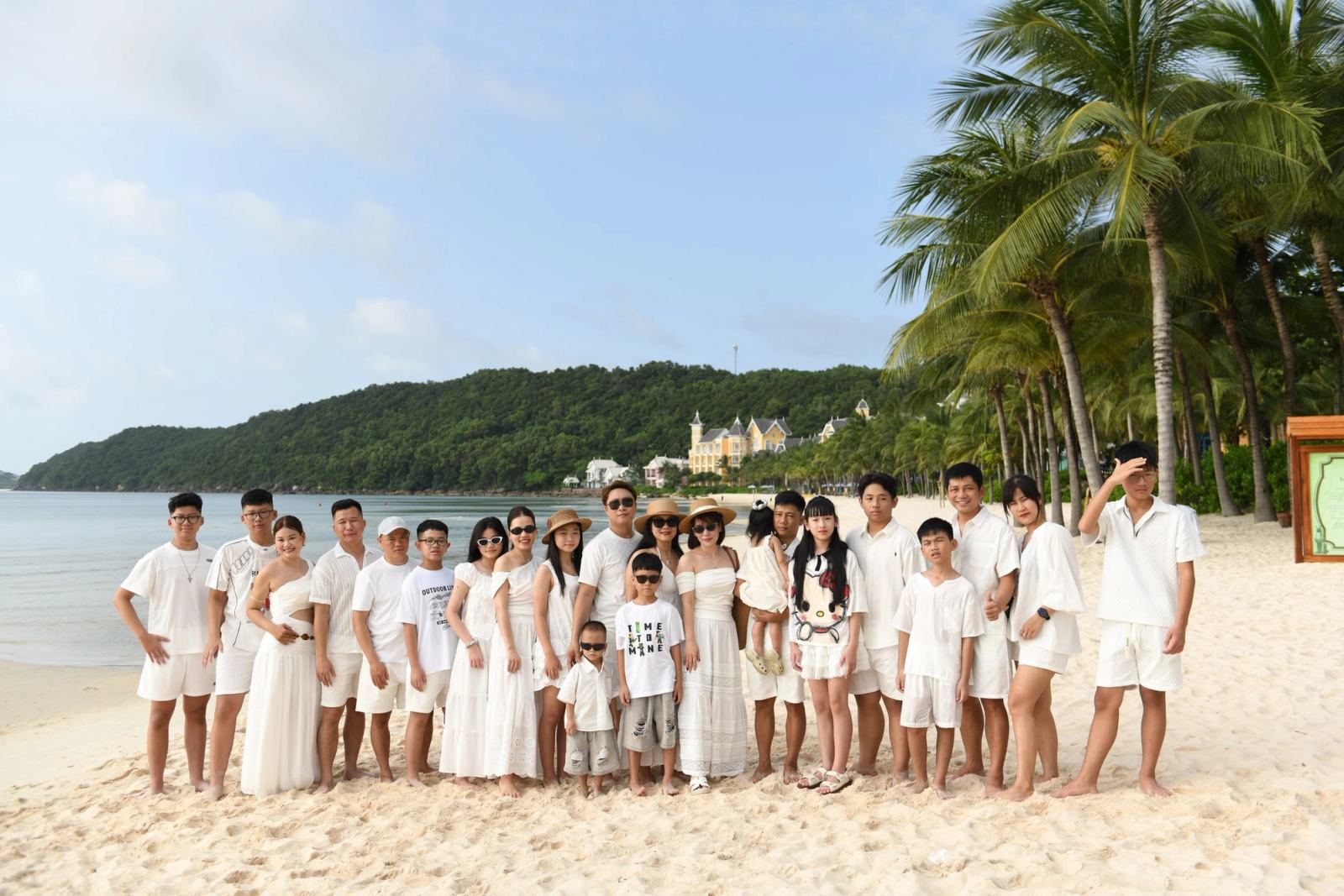 Large family group in white outfits posing on a beach beside palm trees.