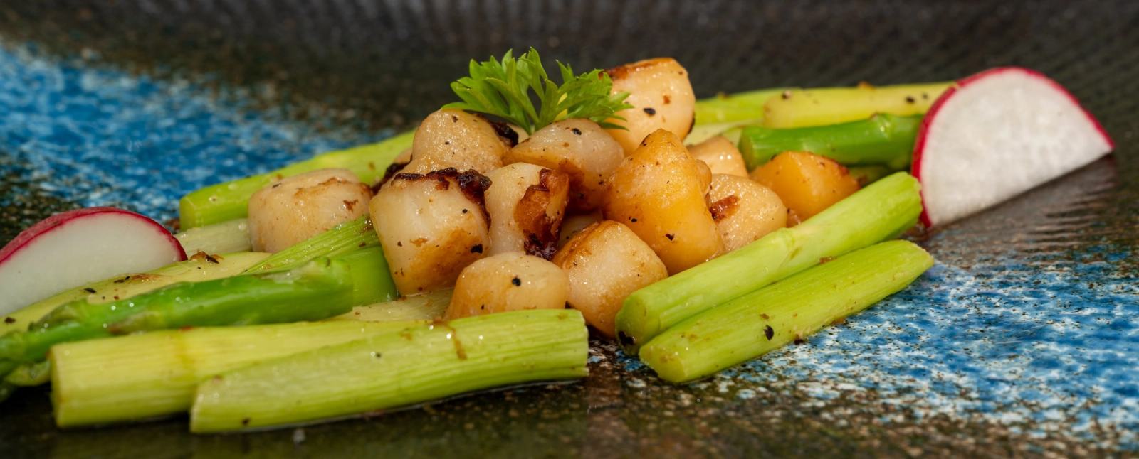 Close-up of asparagus, golden seared pieces and radish on a textured plate.