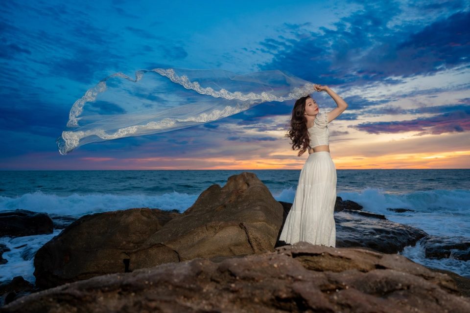 Woman in a white dress holding a flowing scarf above coastal rocks at sunset.