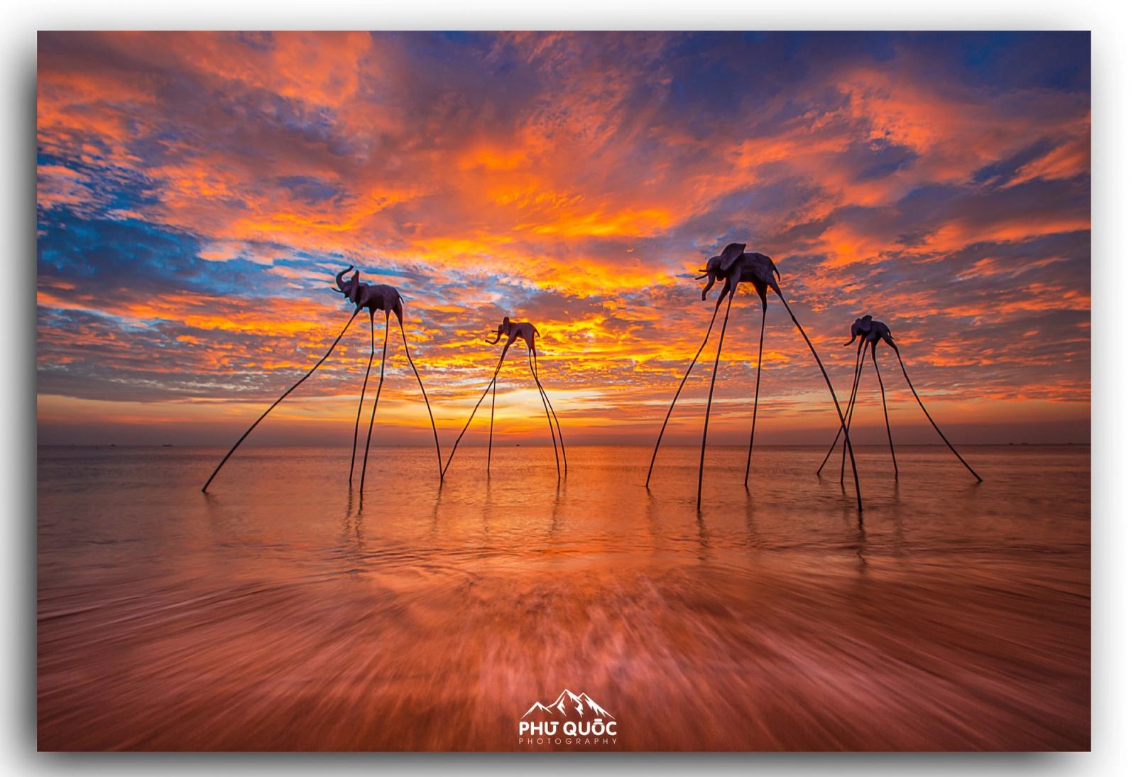 Tall sculptural figures standing in shallow water beneath fiery sunset clouds.