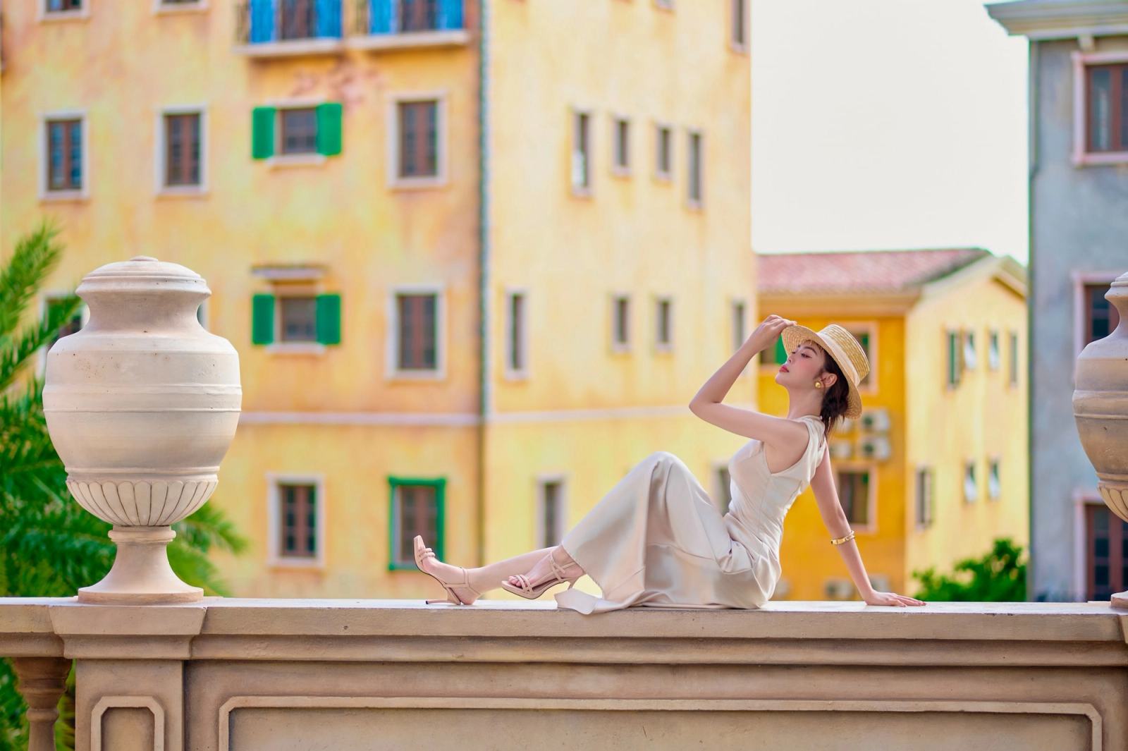 Woman in a sun hat seated on a stone balustrade in front of colourful buildings.