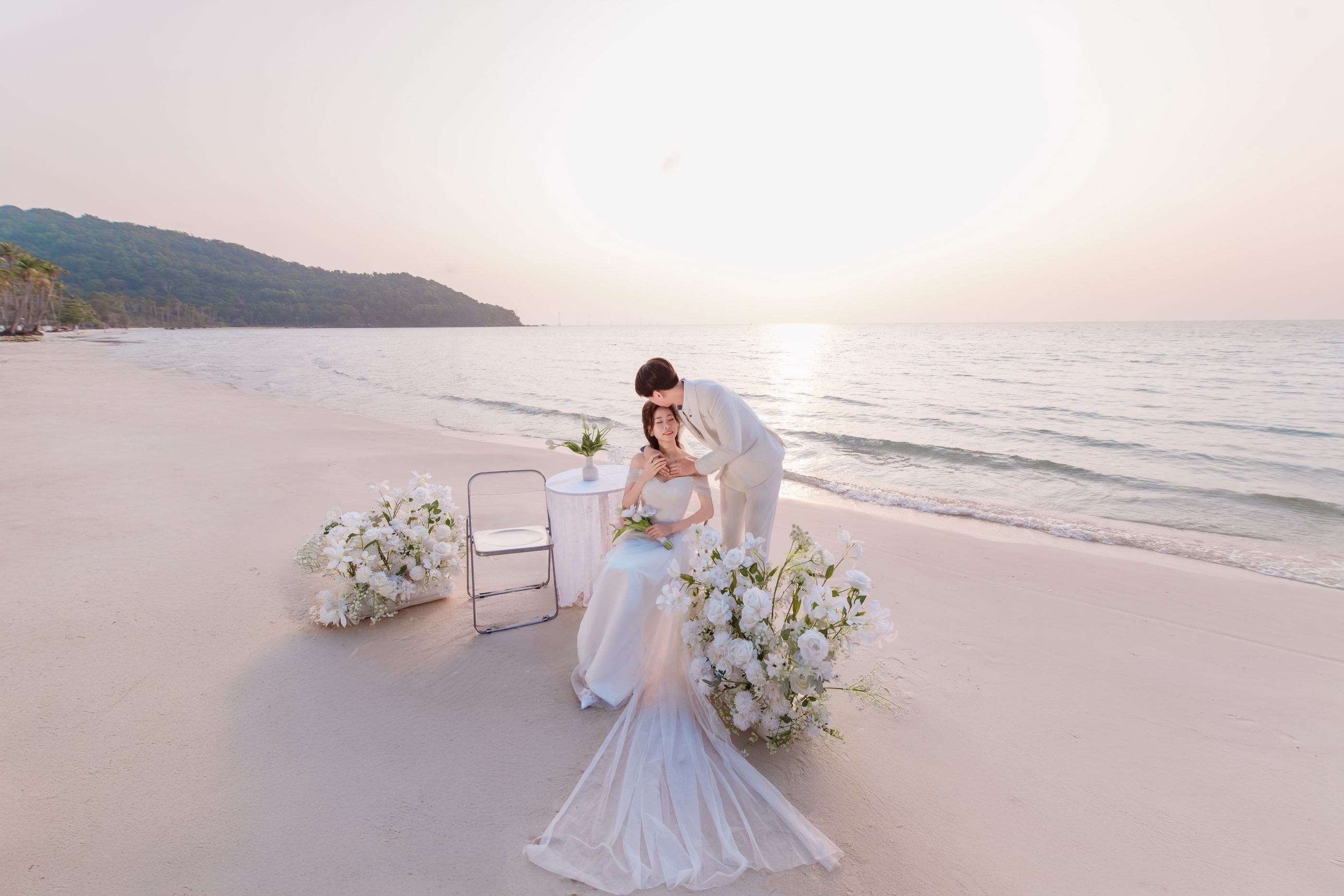 A couple in wedding attire beside flowers on a beach.