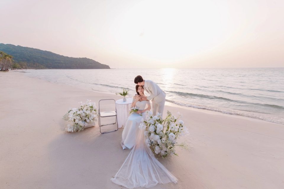 A couple in wedding attire beside flowers on a beach.