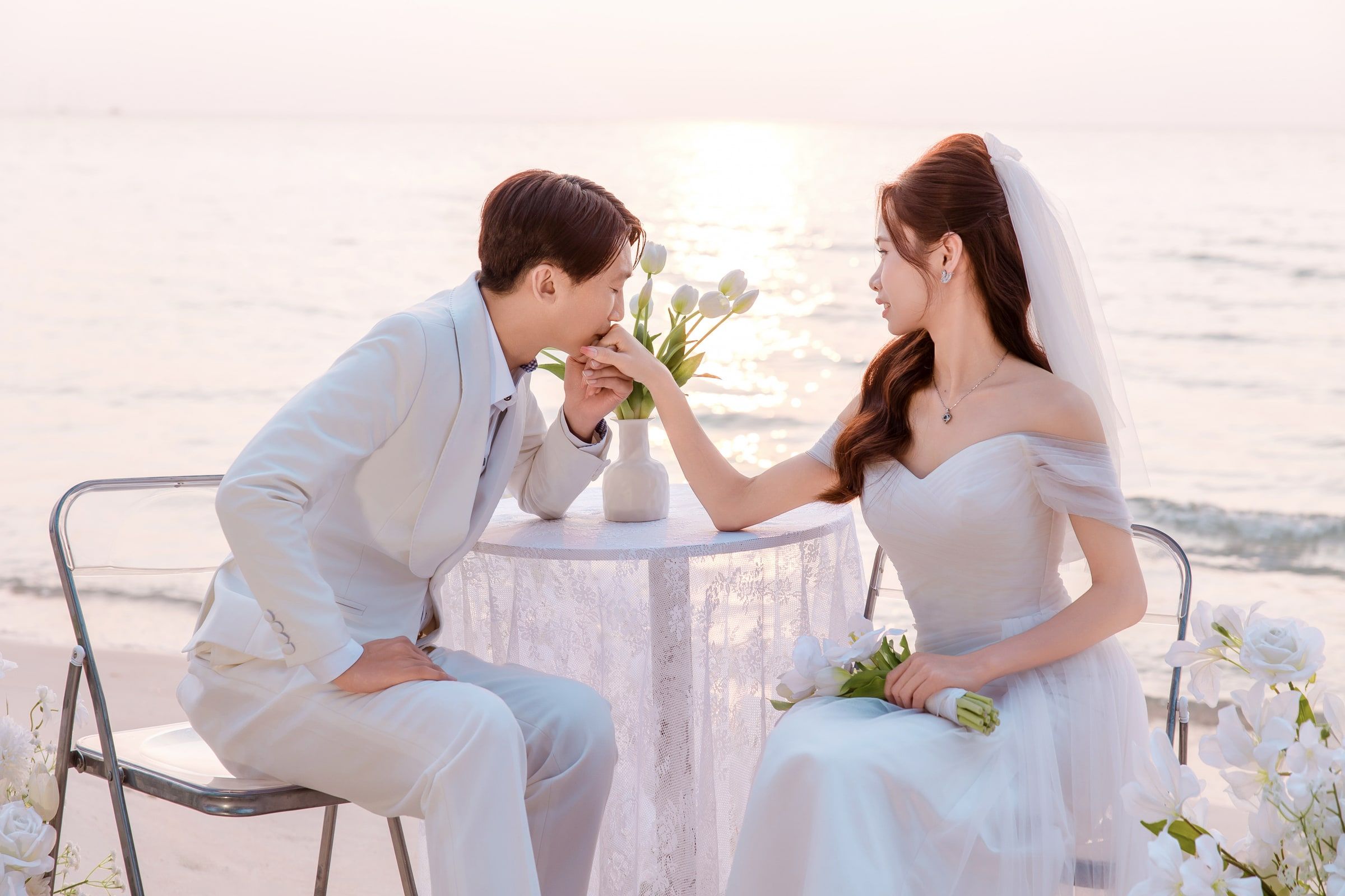 Man kissing his partner's hand across a small lace-covered table with white flowers beside the sea.