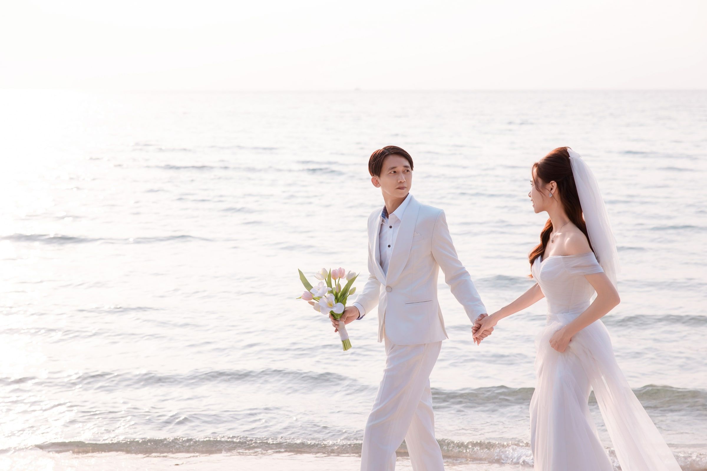 Couple in white wedding attire holding hands at the shoreline, the man carrying a bouquet.