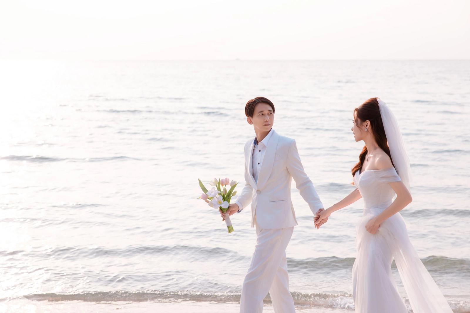 Couple in white wedding attire holding hands at the shoreline, the man carrying a bouquet.