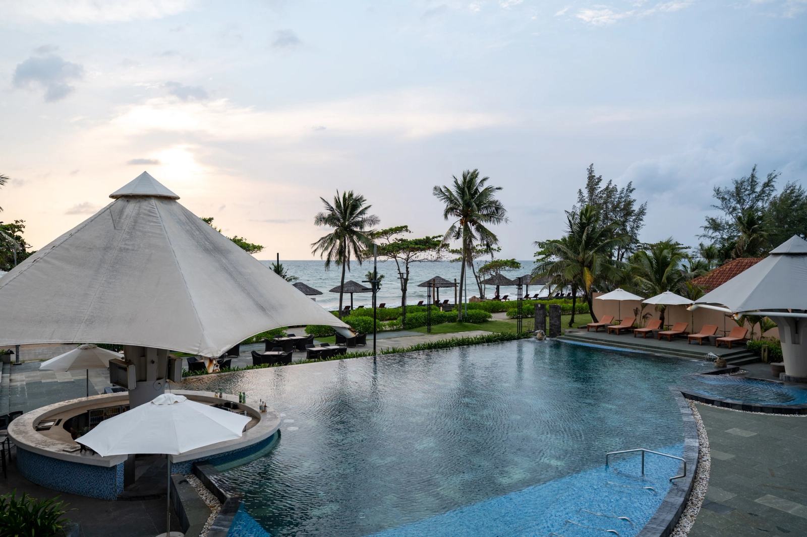 Resort swimming pool with shaded pavilions, palm trees and a view towards the sea.