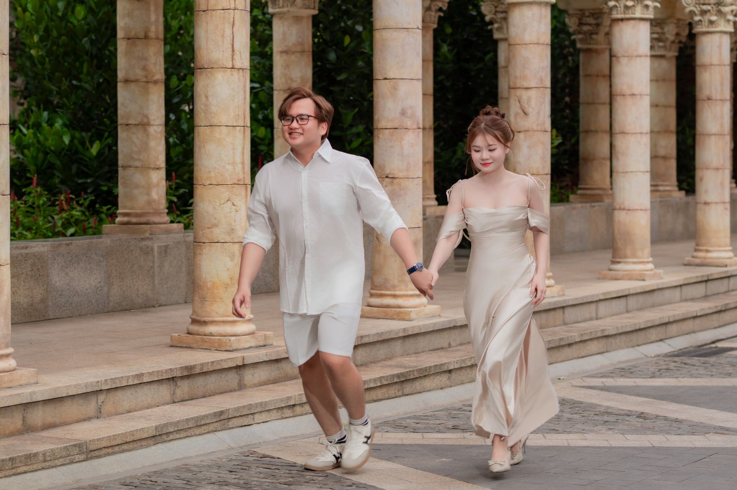 Couple in light outfits walking hand in hand beside a row of stone columns.