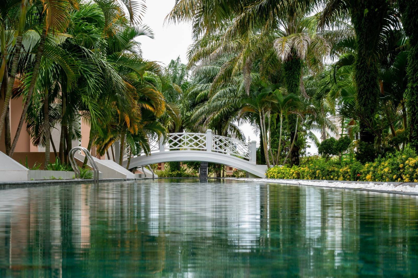 A white footbridge crossing a swimming pool surrounded by dense tropical planting.