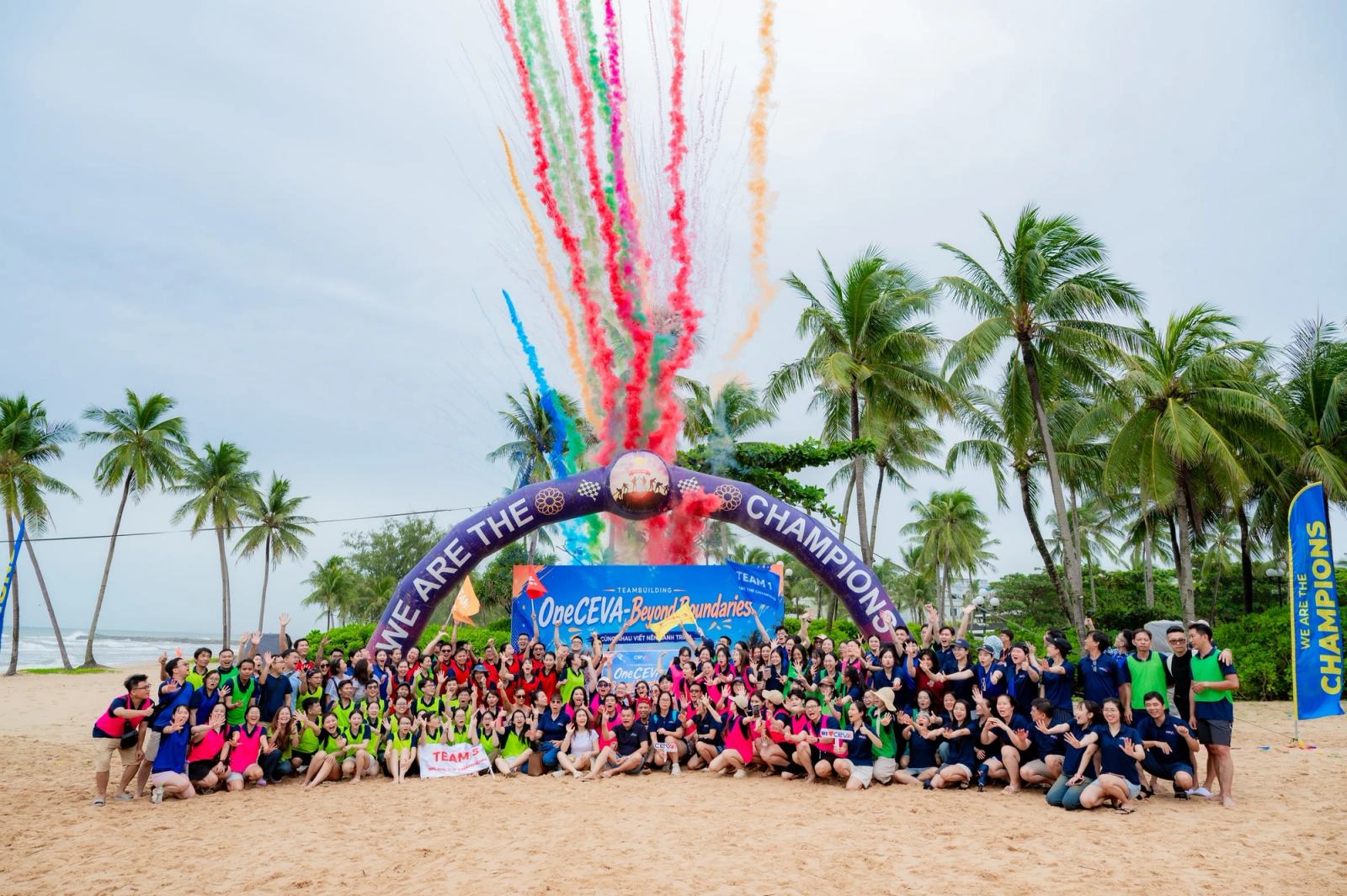 Large group gathered beneath an event arch with coloured smoke above a palm-lined beach.