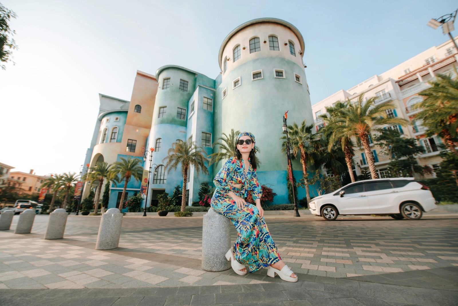 Woman in a patterned dress and sunglasses posing beside pastel-coloured street architecture.