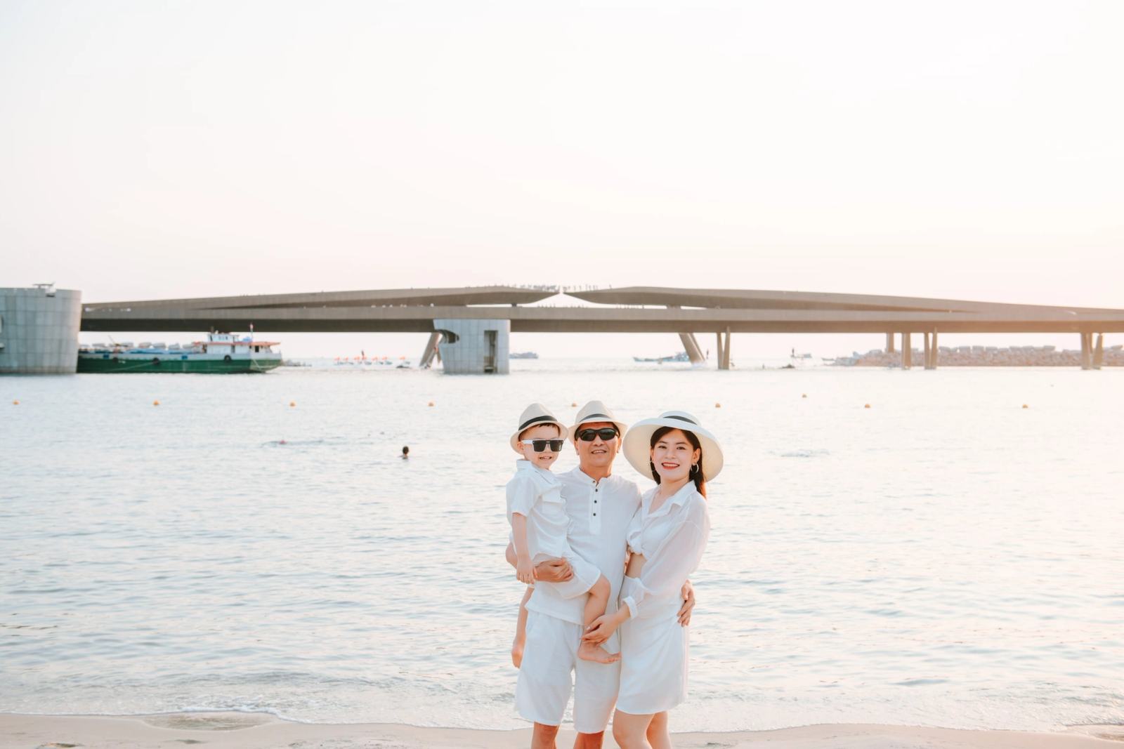 Family of three in white outfits and sun hats posing beside the sea.