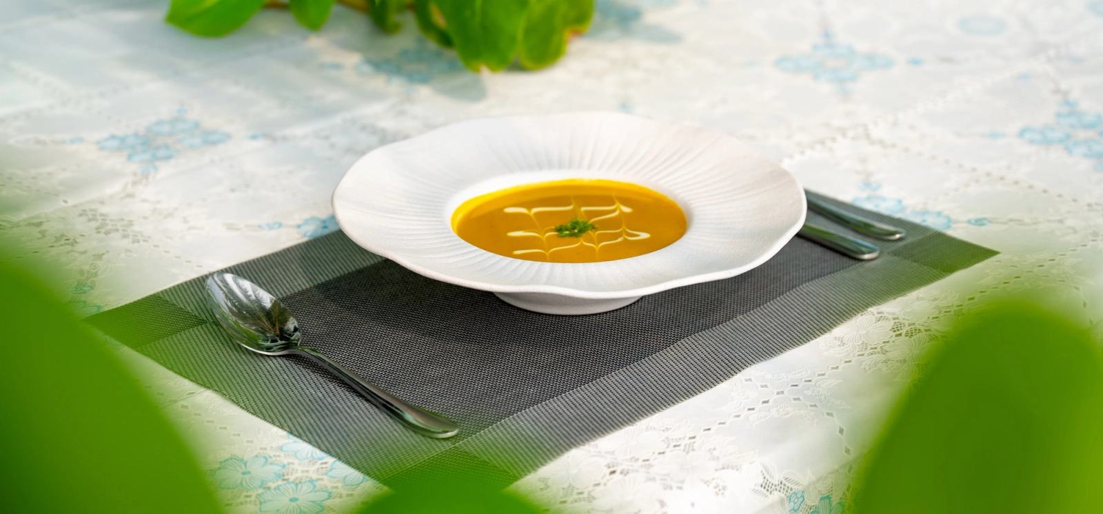 Golden soup in a white bowl, photographed through soft green foliage.