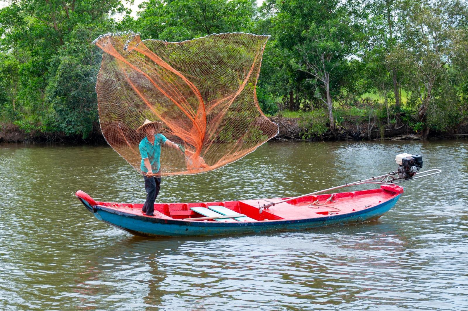 A person casting an orange fishing net from a red boat on a tree-lined river.