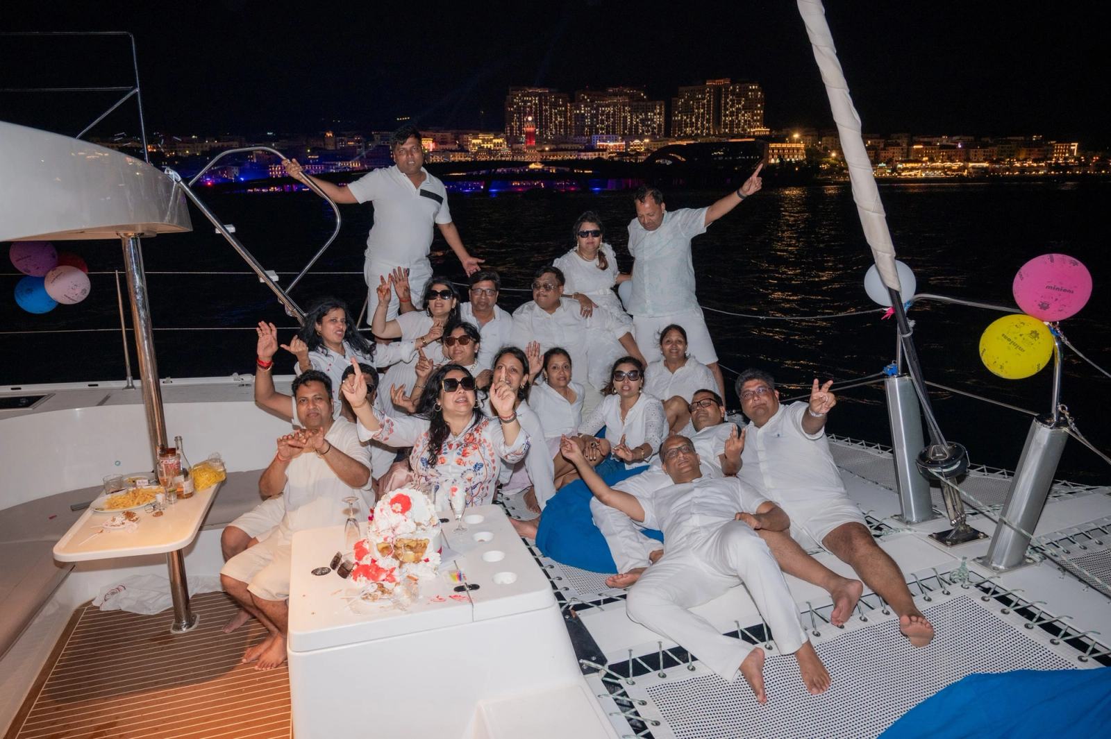 Guests gathered around a cake on a yacht at night with waterfront lights behind them.