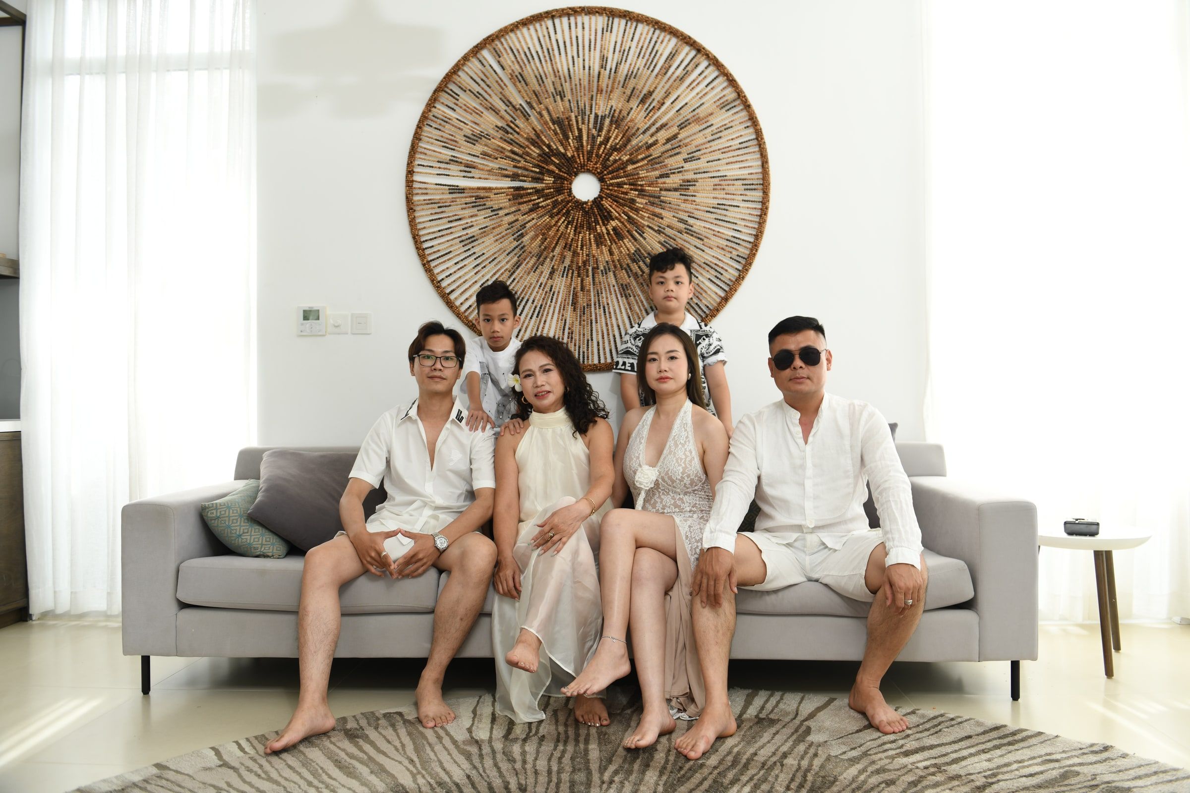 Family in white outfits gathered around a grey sofa beneath a circular wall decoration.