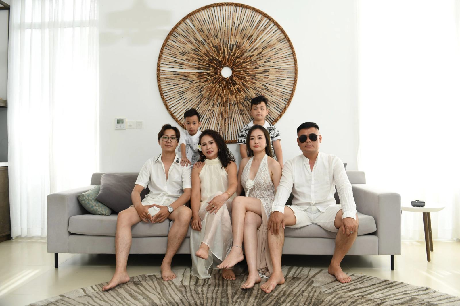 Family in white outfits gathered around a grey sofa beneath a circular wall decoration.