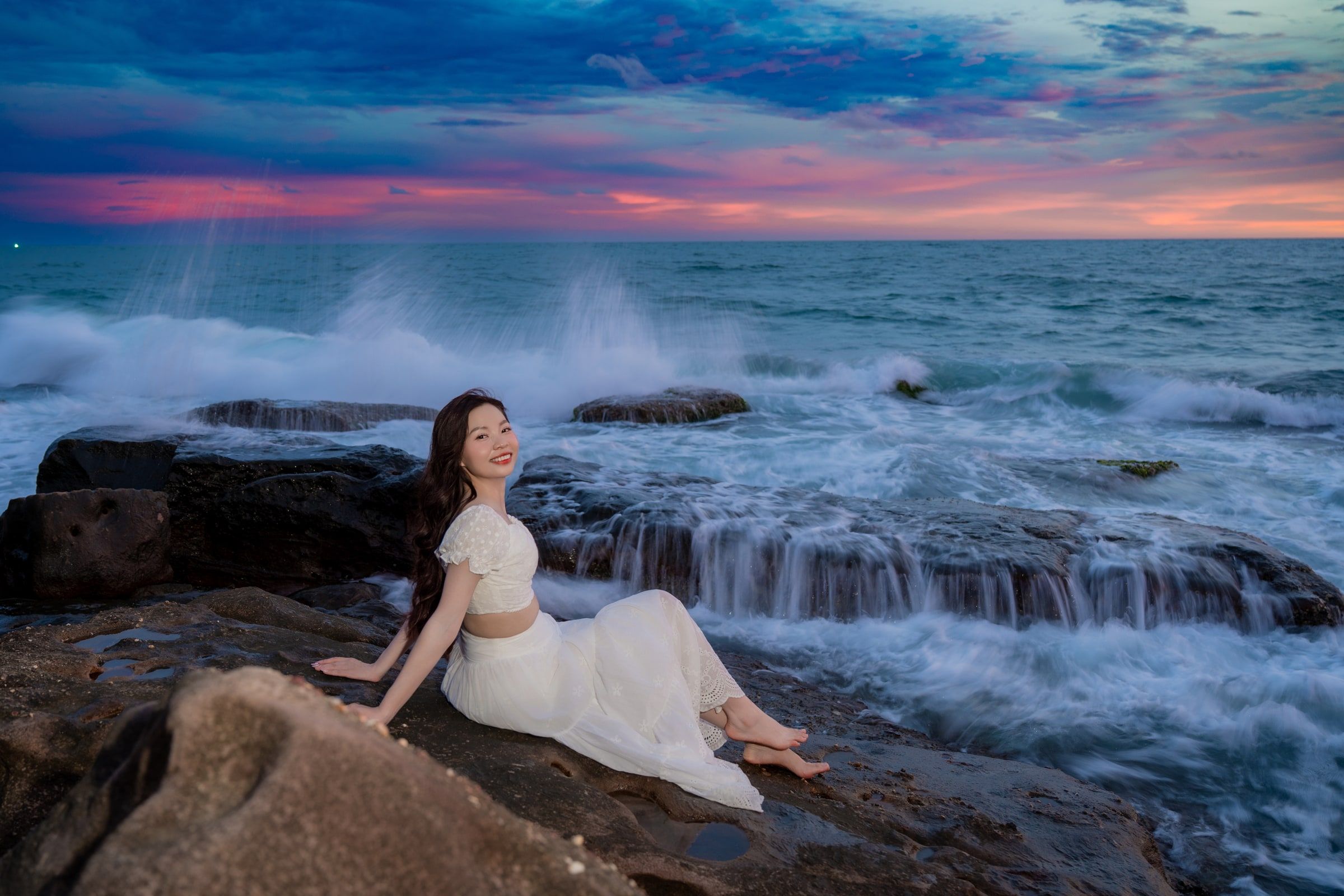 Woman in white seated on coastal rocks as waves break beneath a pink and blue evening sky.