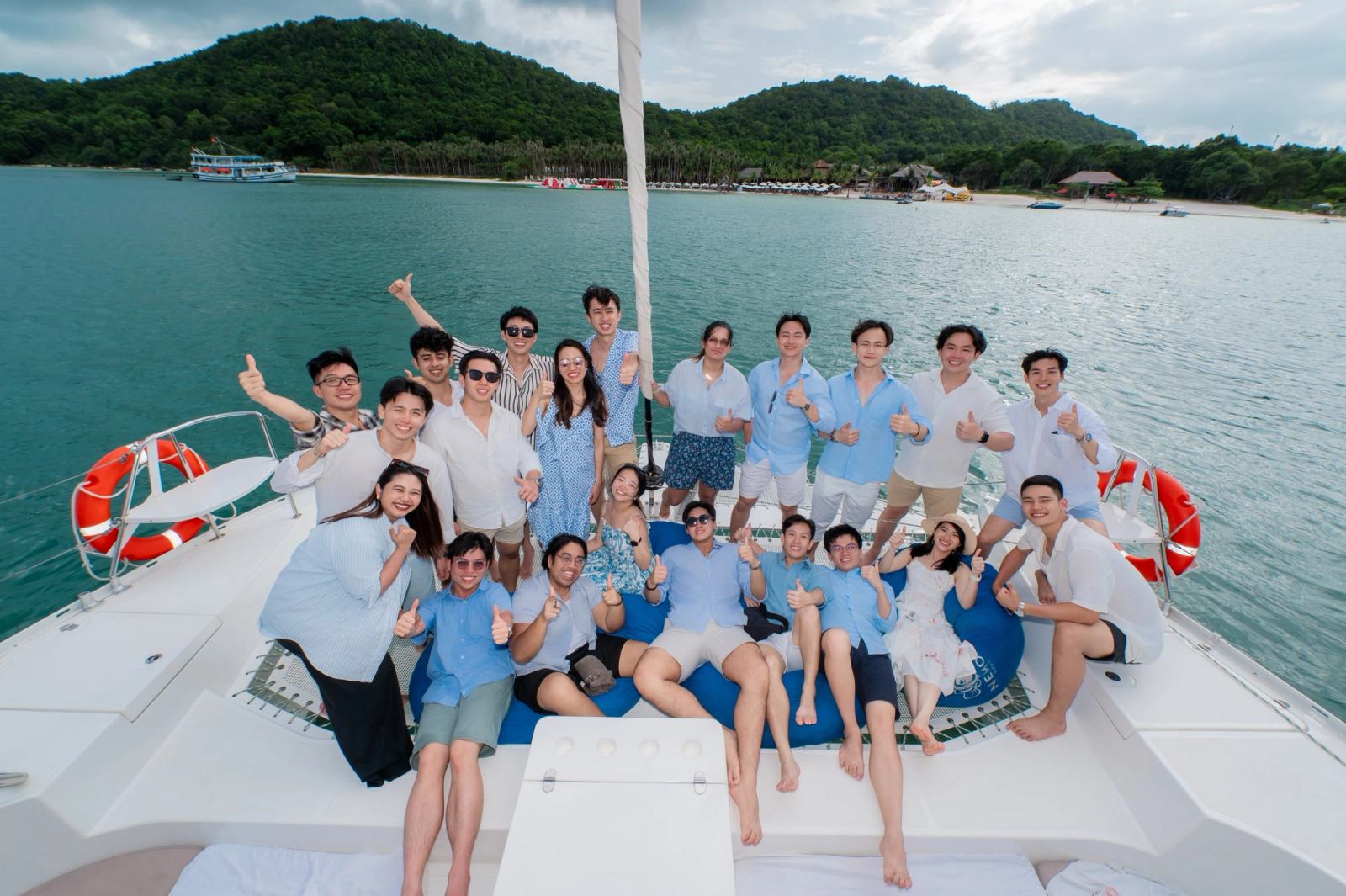 Guests in blue and white posing together on a catamaran beside a green shoreline.