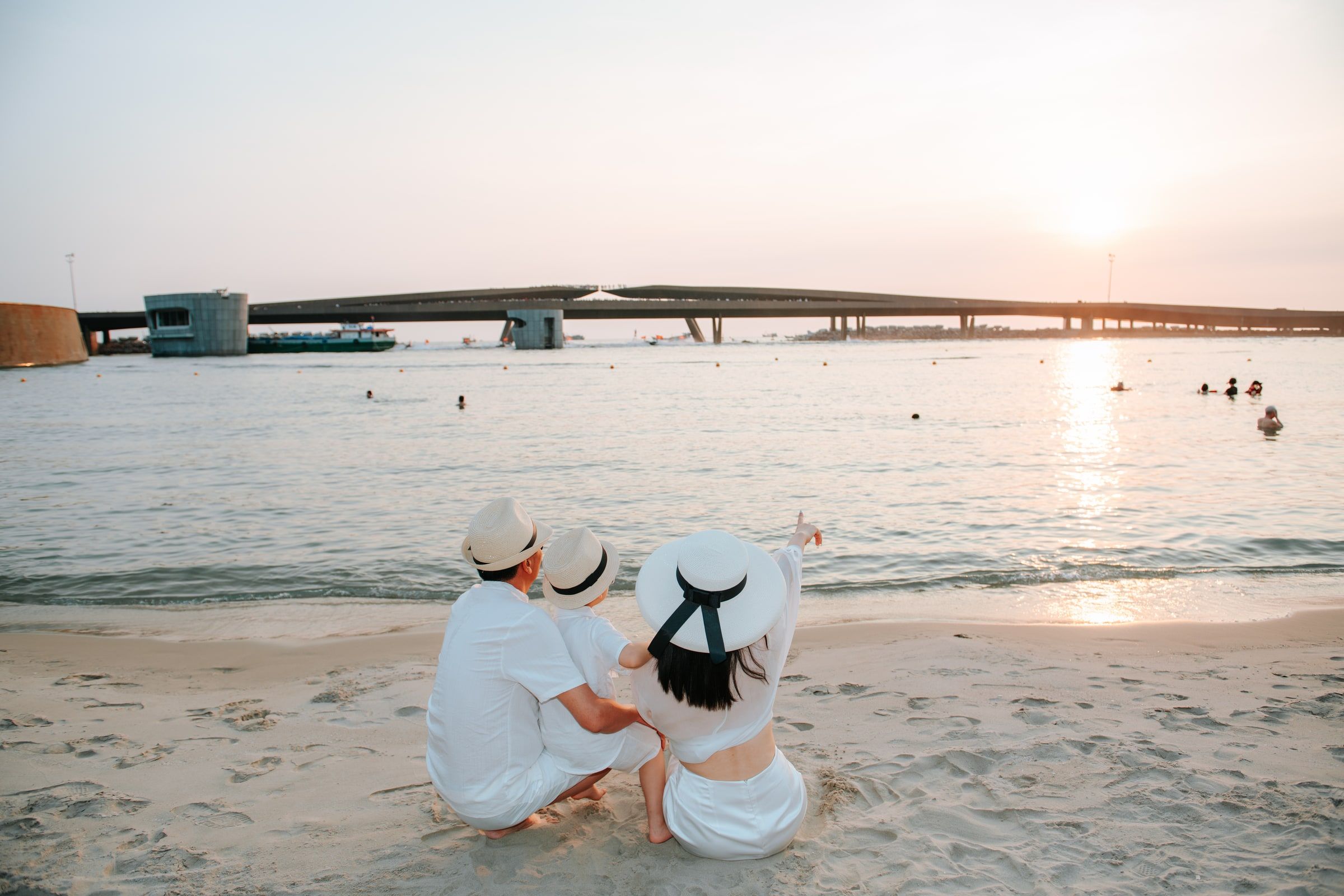 Family seated on the sand looking towards the sea and sunset, seen from behind.