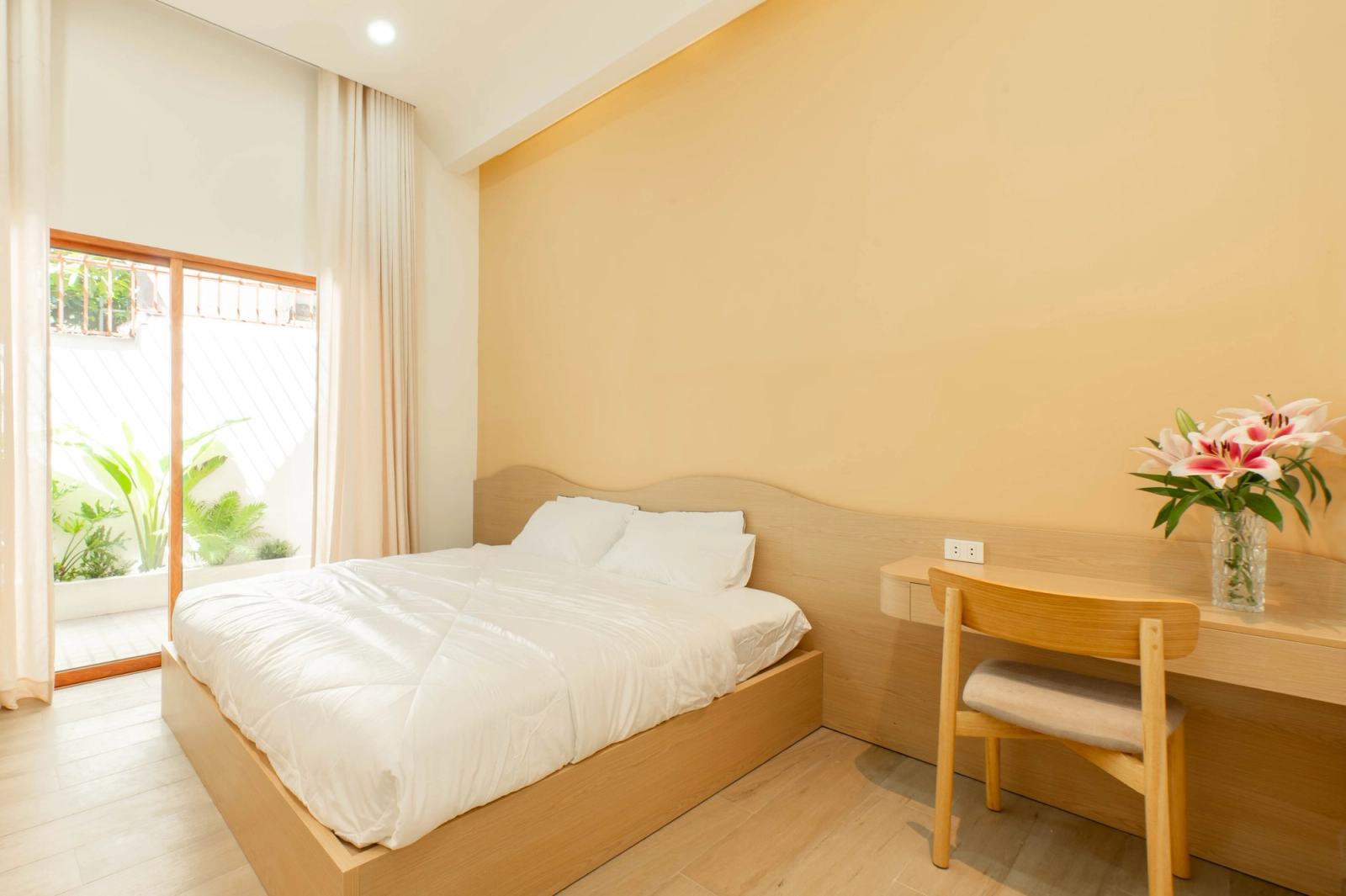 Warm-toned bedroom with a double bed, timber desk and glass doors beside green plants.