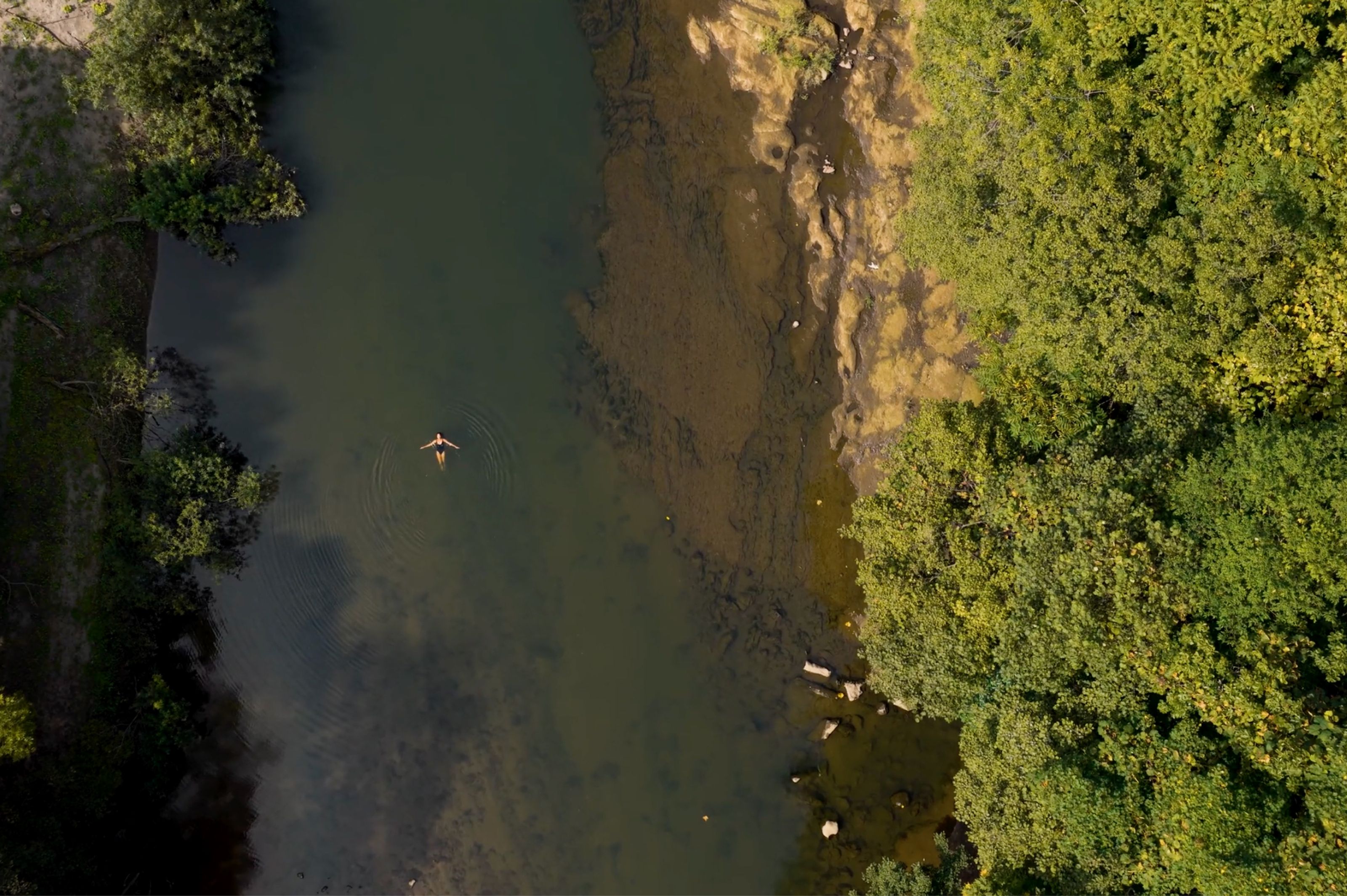 Aerial view of a person floating on their back in a river surrounded by green trees.