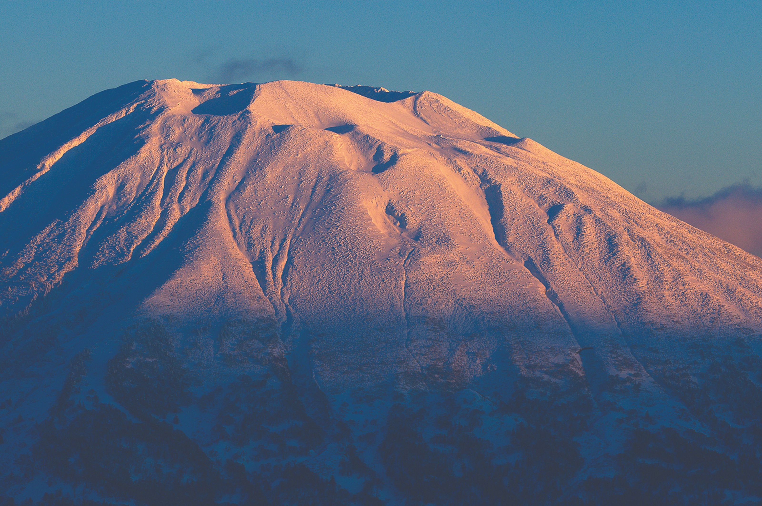 A snow-covered mountain peak, bathed in golden light on its upper slopes with blue shadows below, against a clear sky.