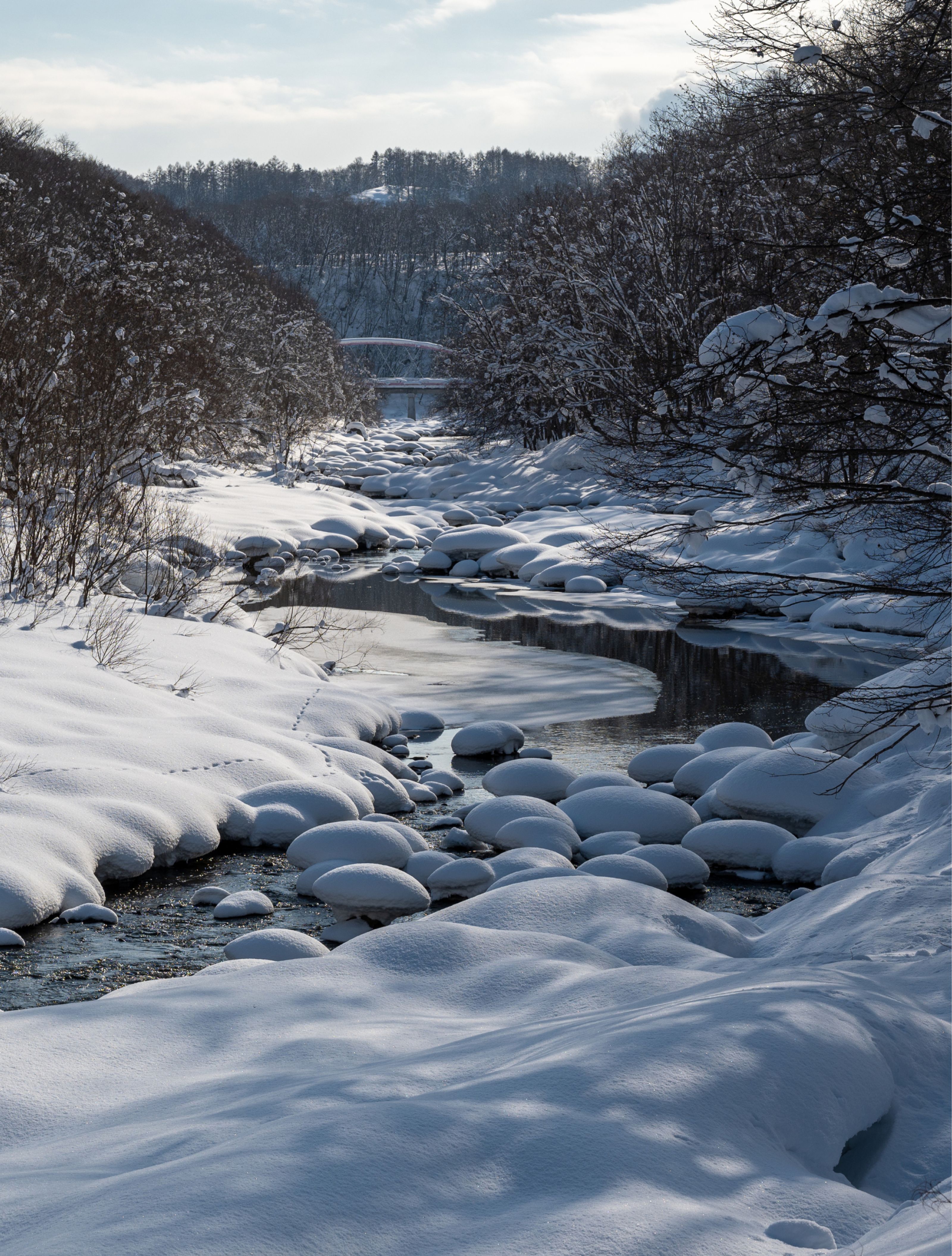 A winding river flows through a snowy winter landscape, with snow-covered rocks and a distant bridge.