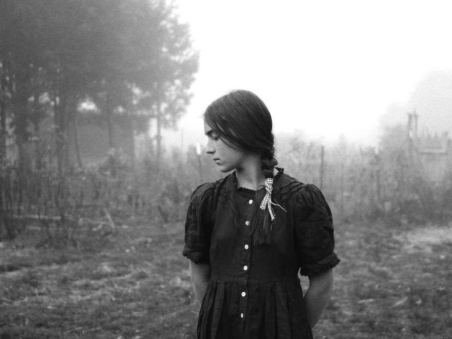 Jeune fille regardant vers la gauche, en extérieur dans une clairière, avec des arbres en arrière-plan à gauche.