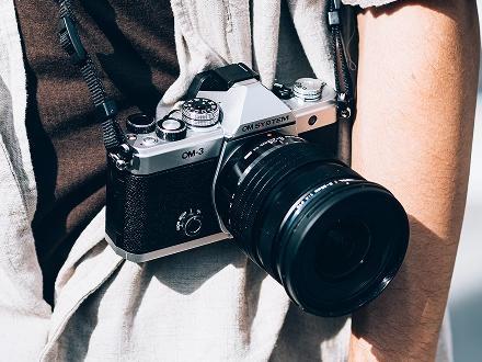 OM SYSTEM OM-3 camera hanging from the neck strap over the should of a person out taking photos.