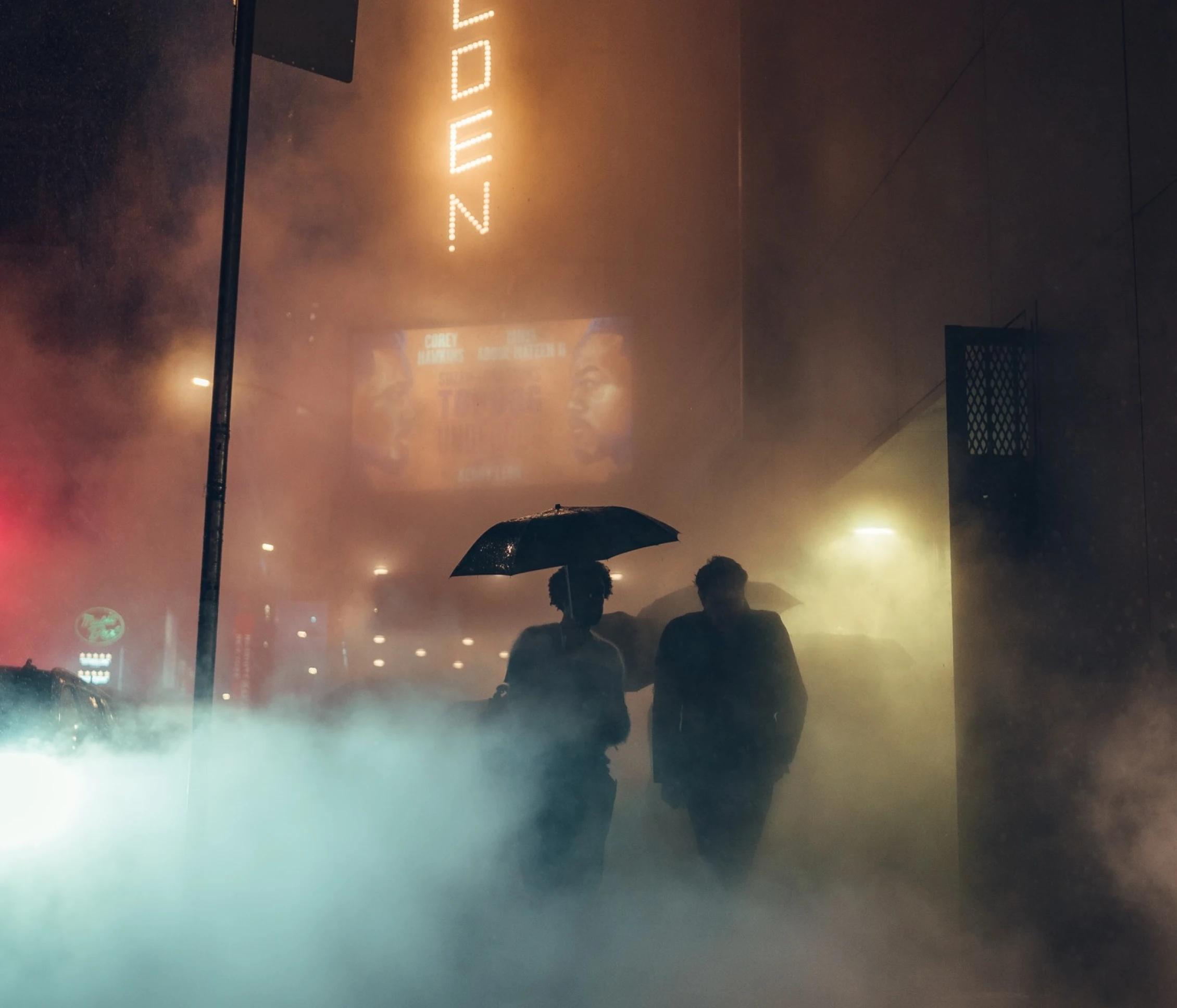 Foggy night in the city with a couple walking on the sidewalks with the girl having an umbrella in her hand. 