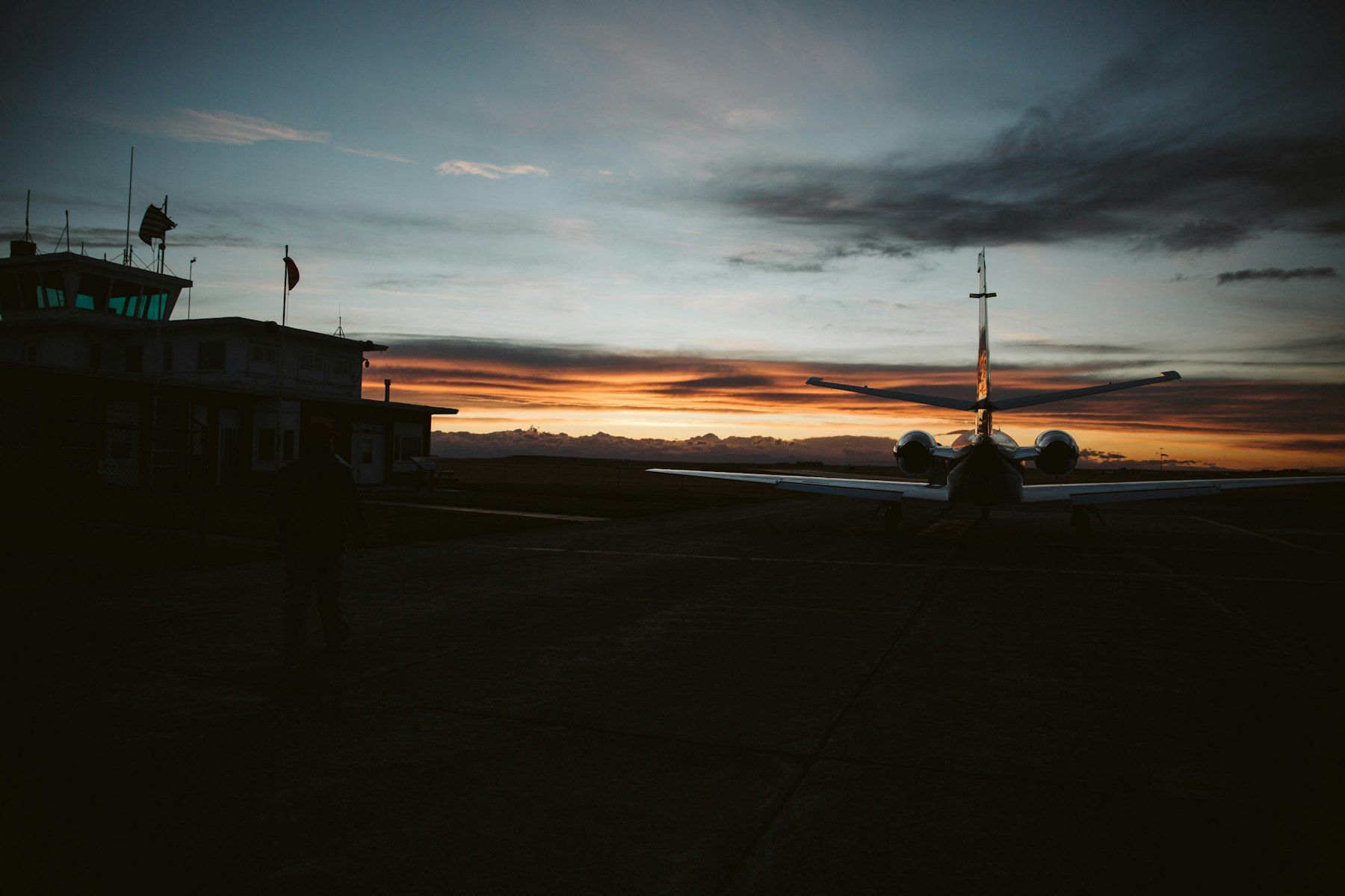 Silhouetted person crossing an airport apron at dusk toward the terminal and control tower, a business jet parked against the sunset