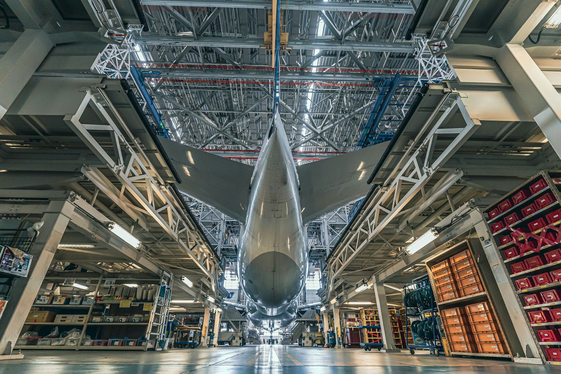 Rear view of a widebody airliner parked in a maintenance dock, its tail rising into the hangar roof, parts shelving and tool carts on both sides