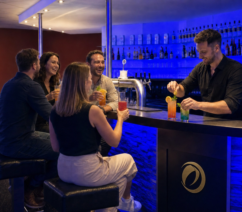 A bartender garnishes drinks for four guests at a blue-lit bar.