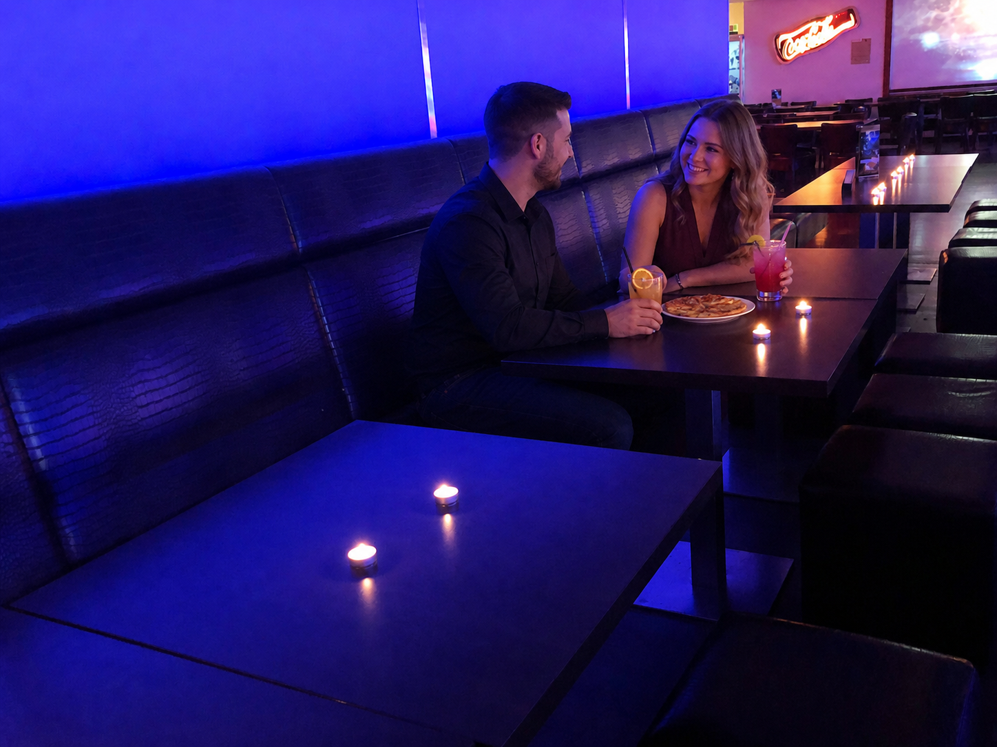 An adult couple sits with drinks and pizza in the blue-lit lounge.