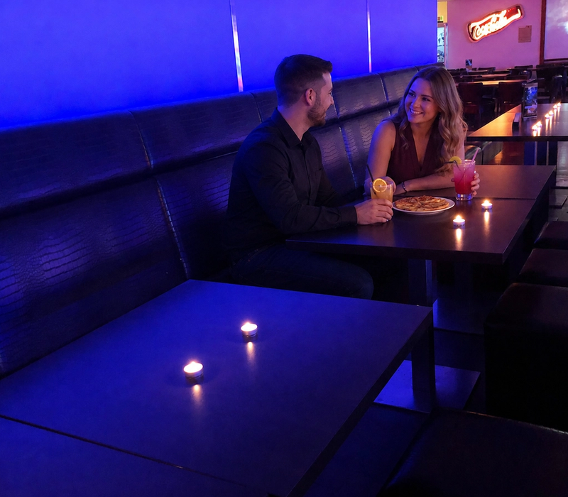 An adult couple sits with drinks and pizza in the blue-lit lounge.