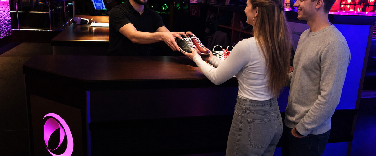 A man hands bowling shoes to two adult guests at the counter.