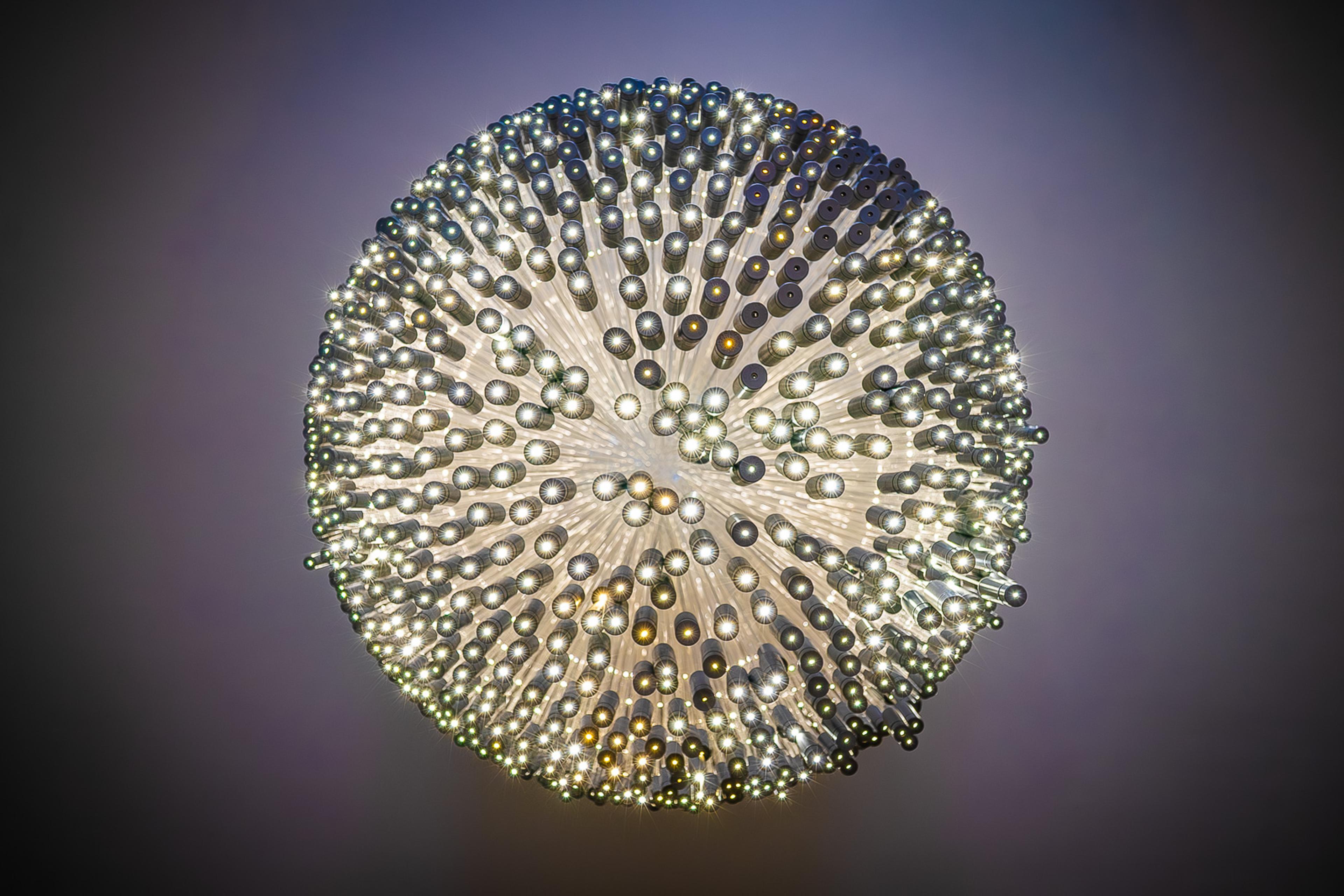 A large spherical chandelier suspended against a dark ceiling in Paris, its surface dense with small light sources. Photographed from directly below, the glowing globe holds its own gravity - self-contained and impossible to ignore.
