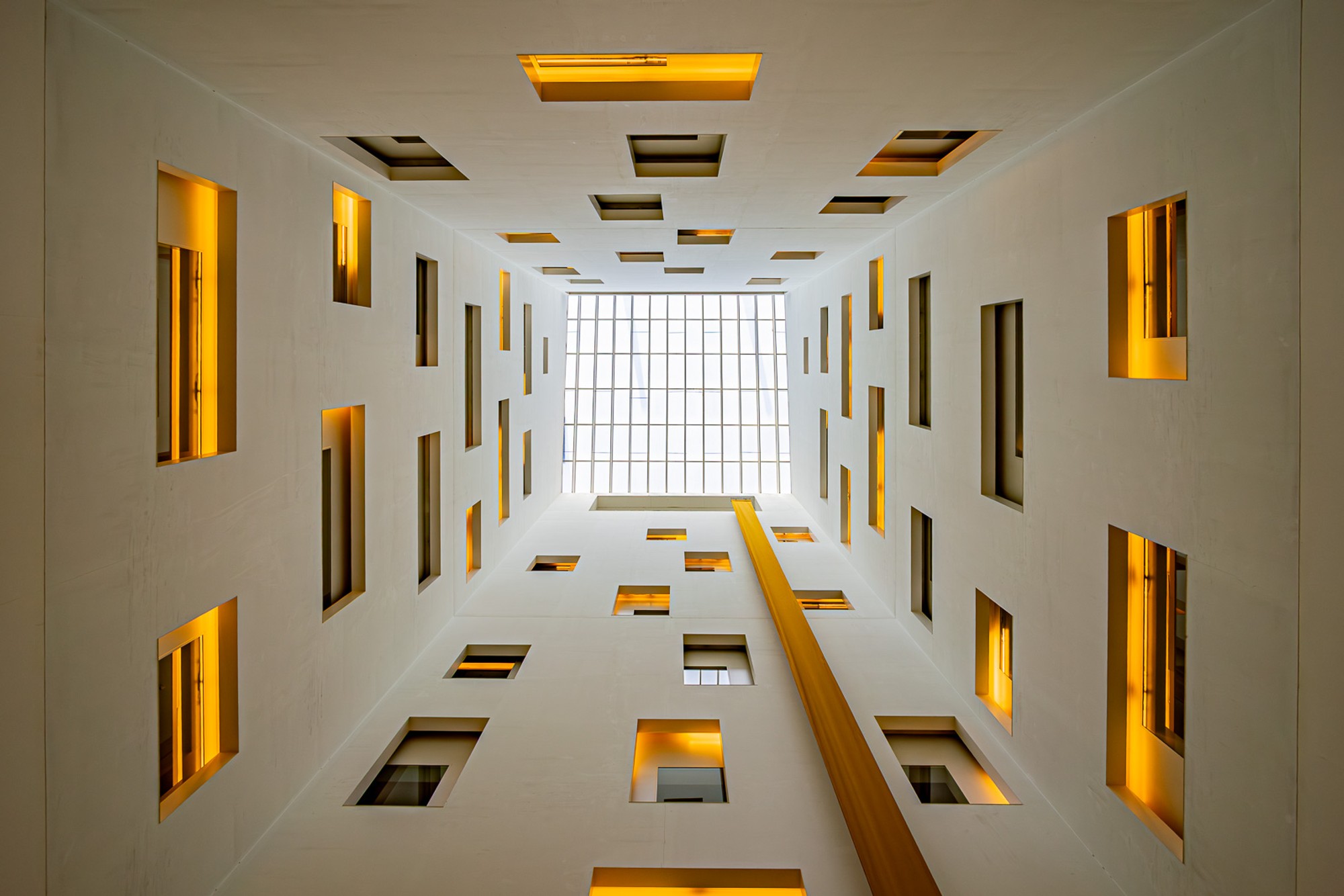 A Barcelona hotel atrium seen from below, its open floors rising in warm amber light. Railings and architectural details recede in perspective toward the ceiling - the kind of interior that makes a case for staying in rather than going out.