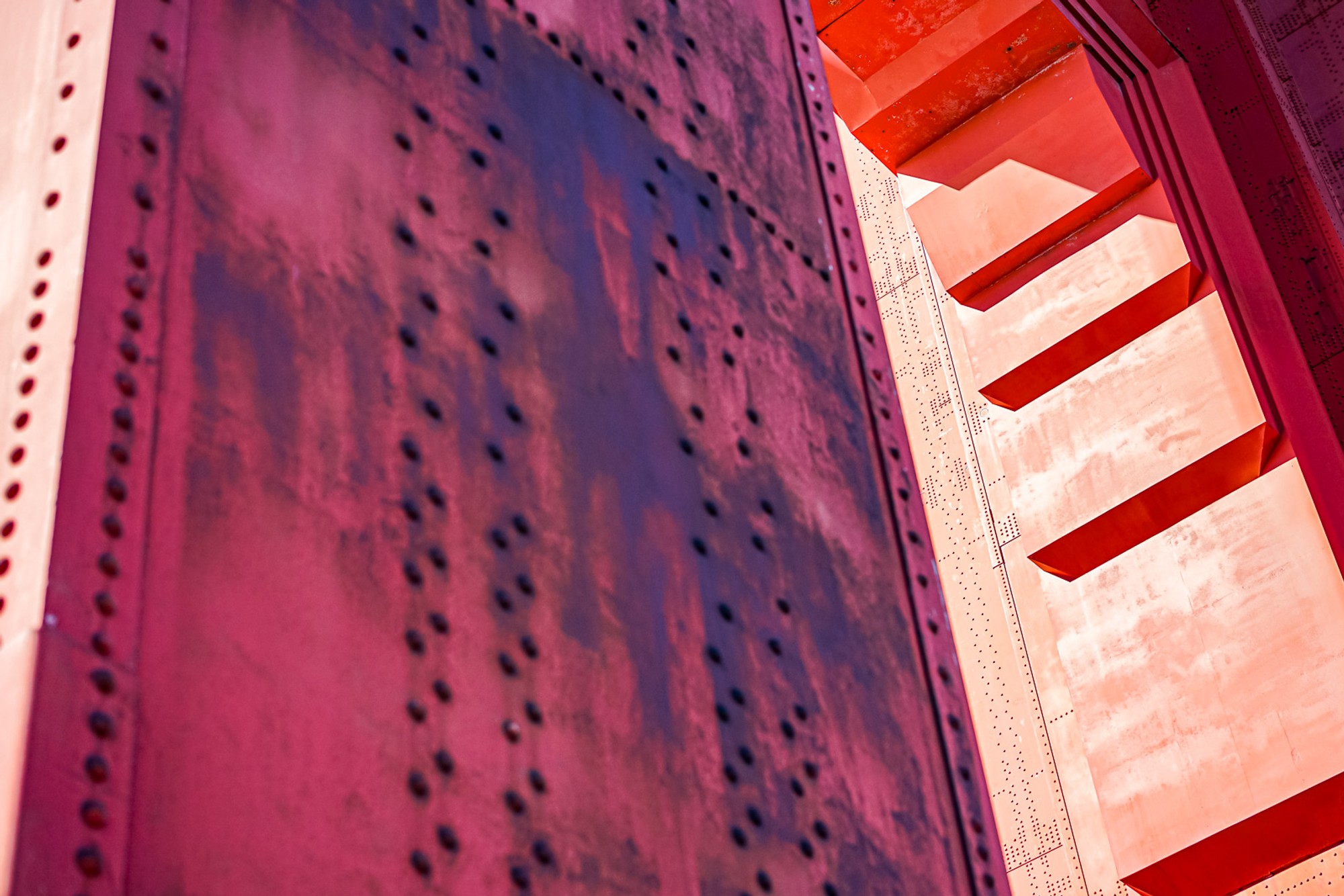 The painted steel hardware of the Golden Gate Bridge photographed close - large rivets and structural members, the orange-red paint worn at the edges. The bridge at the scale of the hand that tightened each bolt.