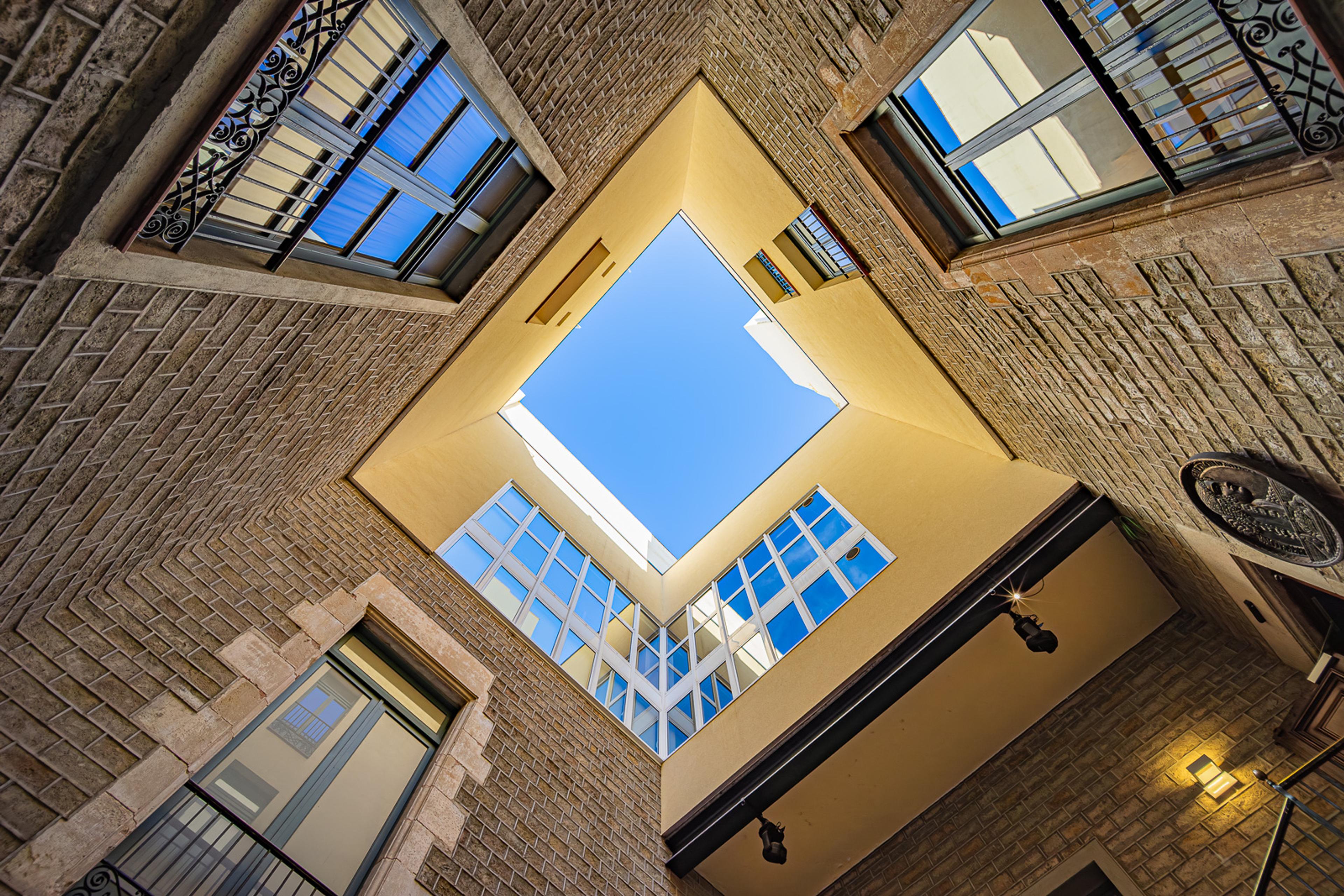 A courtyard in Barcelona's Gothic Quarter shot looking straight up. Aged stone walls and iron balconies frame a precise rectangle of blue sky - the city compressed into four walls and whatever exists above them.