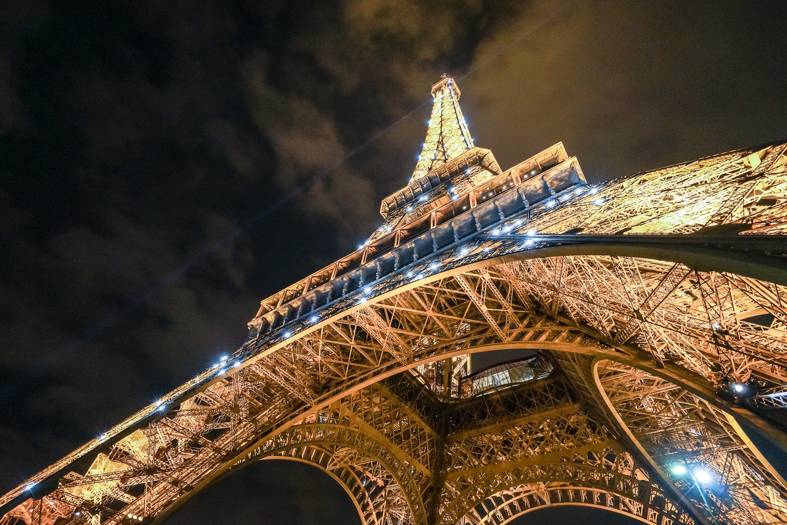The Eiffel Tower photographed from directly below at night, looking straight up its iron lattice toward the illuminated tip. The structure converges on a single bright point - Paris reduced to its most fundamental vertical.