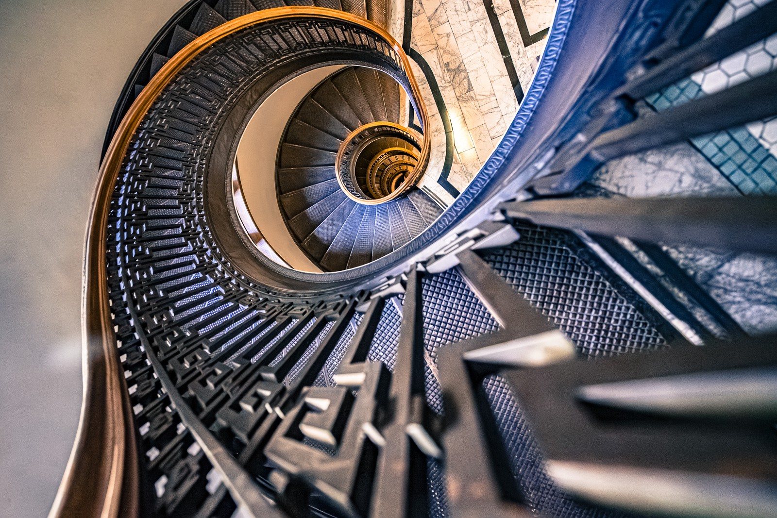 A spiral staircase photographed from directly above, the stairs winding downward in an elegant helix. Each level visible within the spiral - a descent turned into geometry.