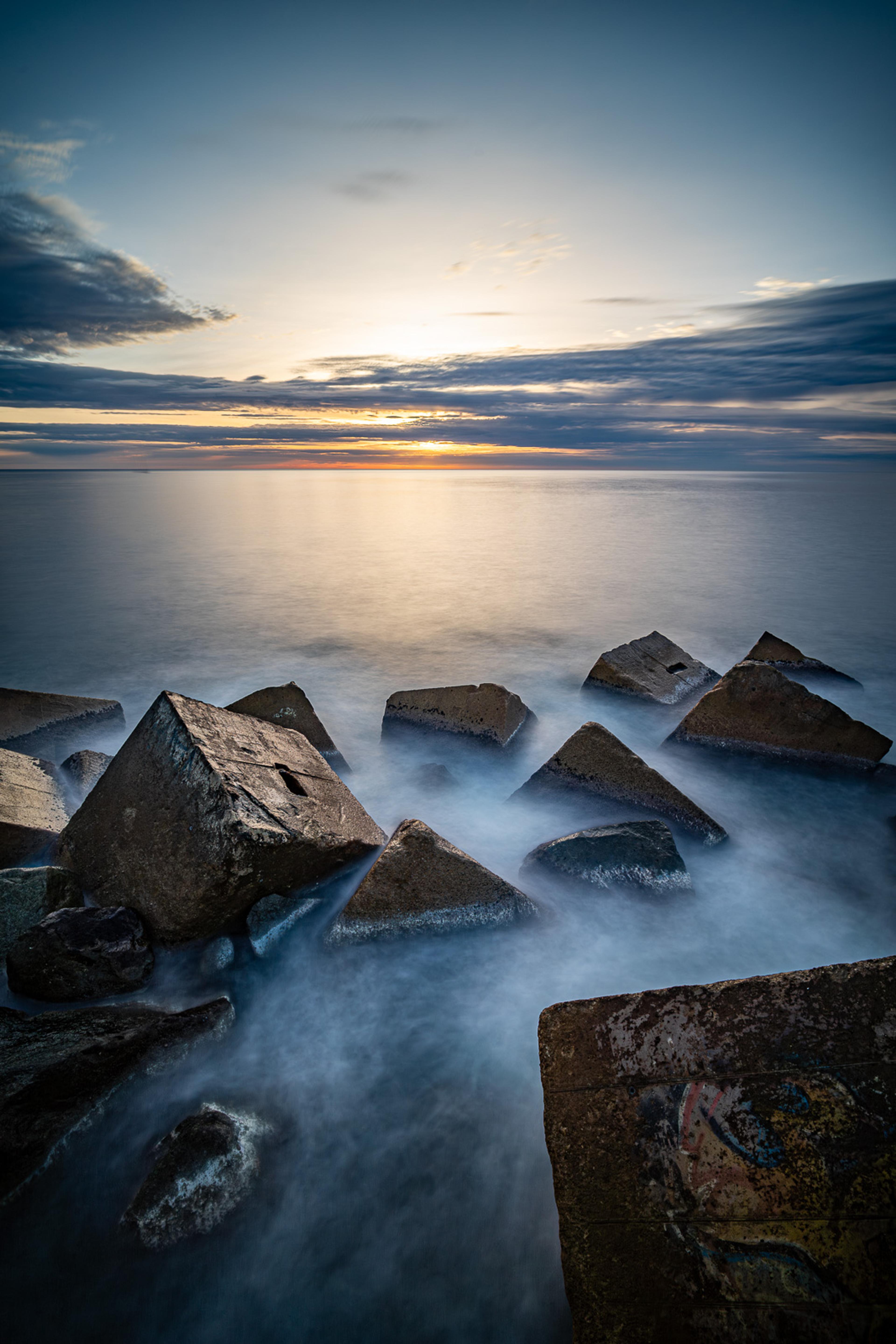 The Barcelona coastline at dawn as a long exposure, dark rocks sitting still while the sea moves around them in soft white trails. The horizon glows with the first light